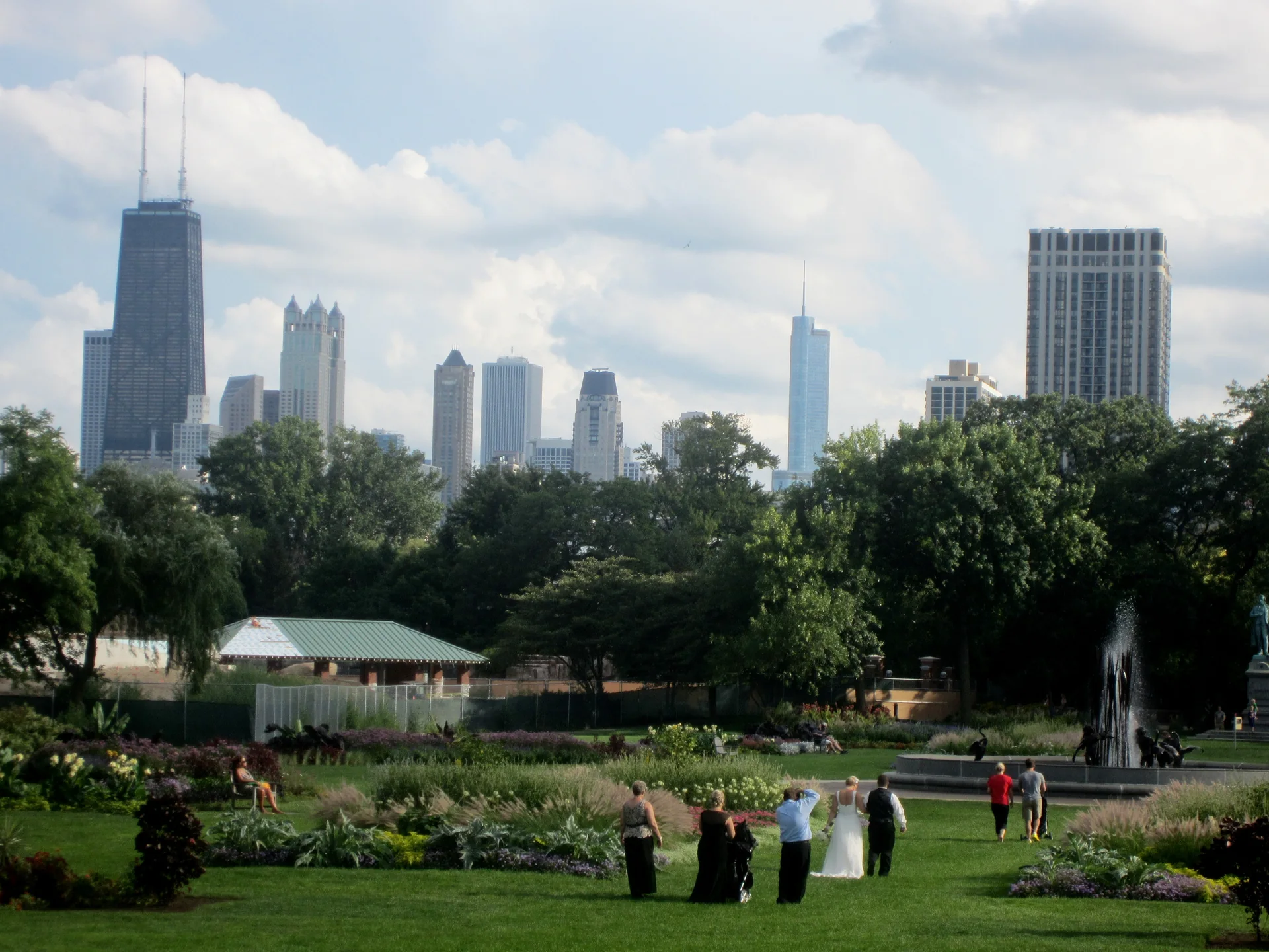 Jardins de Lincoln Park com fonte, árvores, visitantes ao longe e o skyline de Chicago sob céu parcialmente nublado.