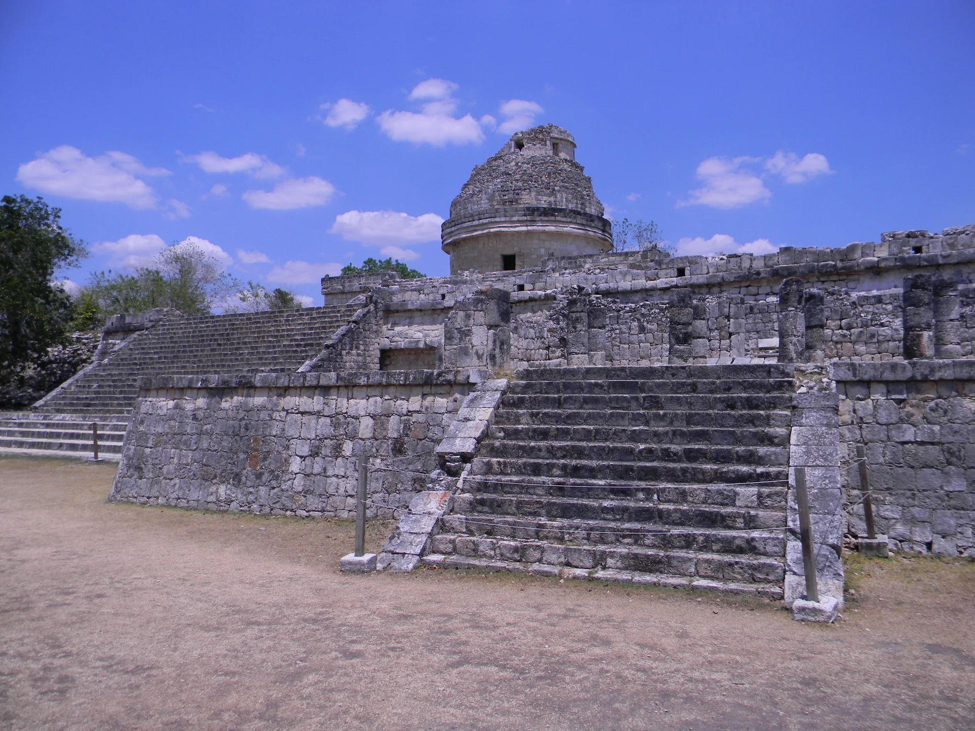 Observatório El Caracol em Chichén Itzá visto atrás de escadarias e cordas de proteção, sob céu azul com nuvens.