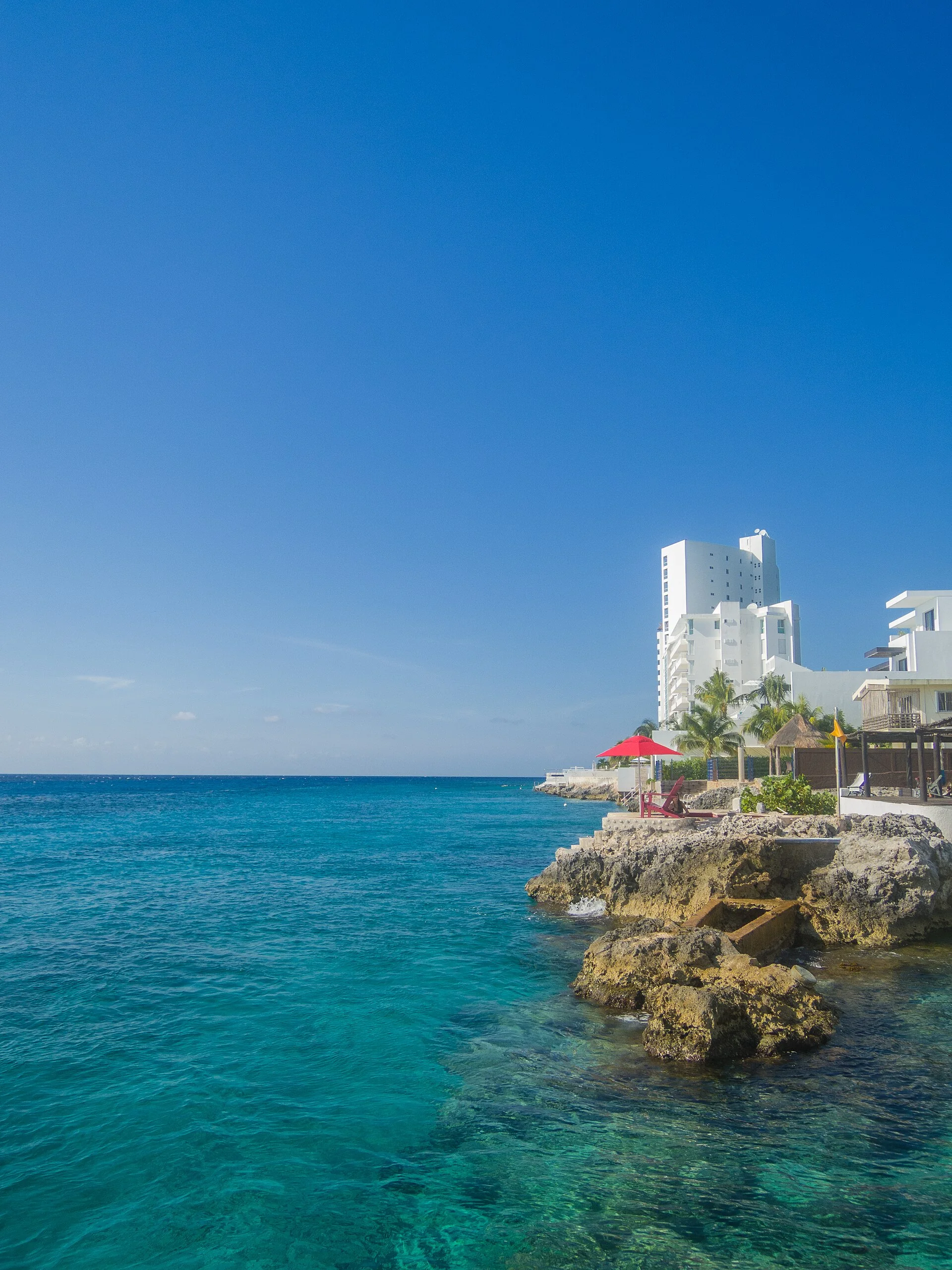 Litoral rochoso de Cozumel com água turquesa transparente e edifícios ao fundo.
