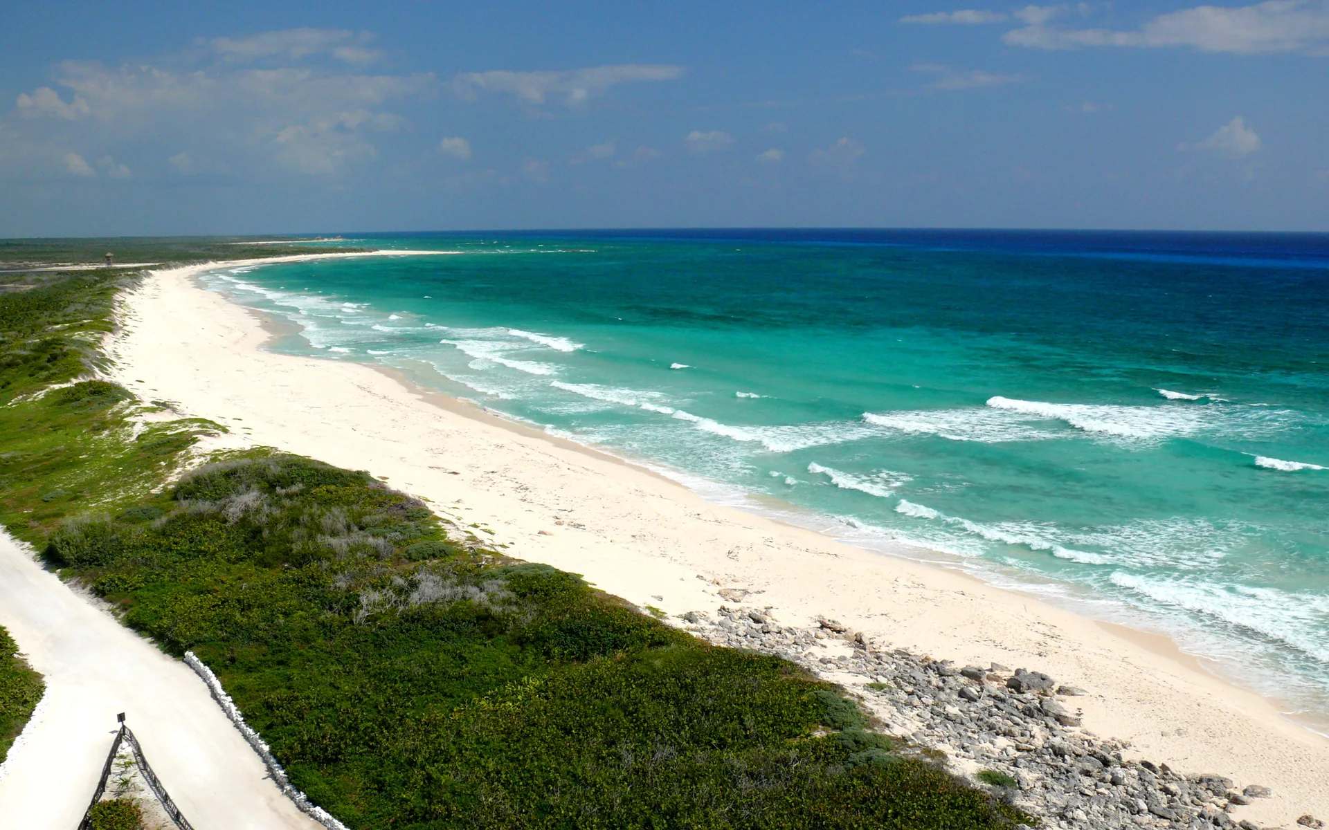 Vista ampla e ensolarada da costa leste de Cozumel, com praia clara, vegetação baixa e mar do Caribe em tons de azul e turquesa.