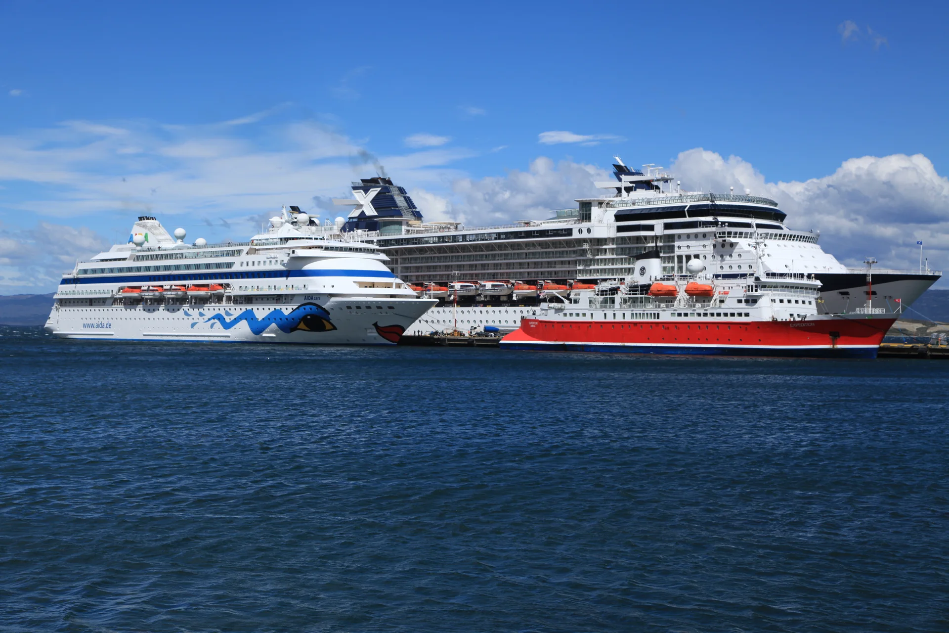 Navios de expedição atracados em Ushuaia, com instalações portuárias e montanhas ao fundo.