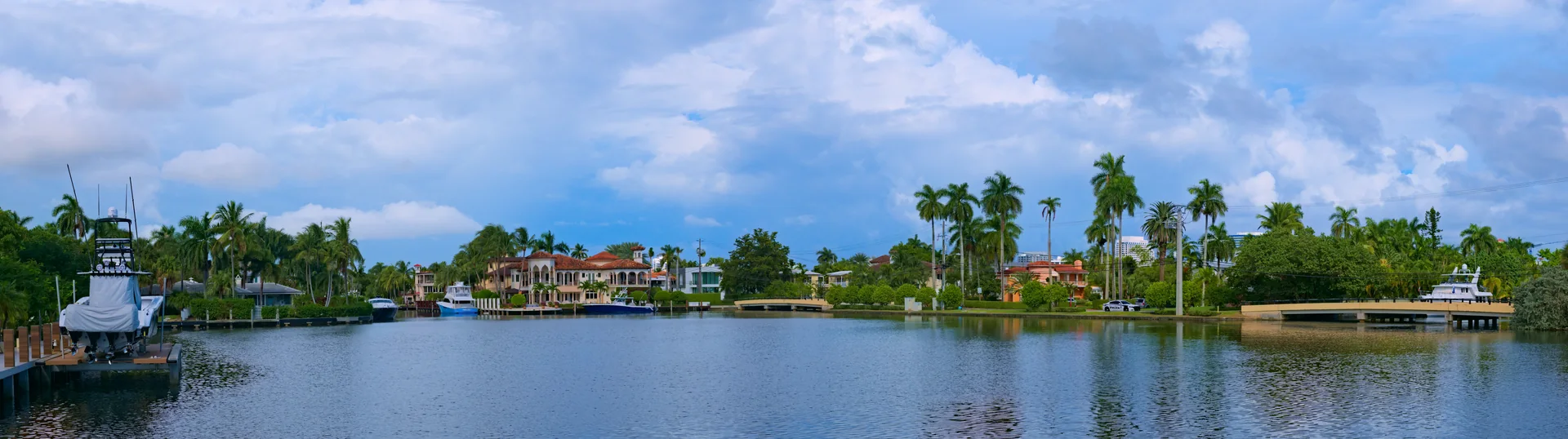 Canal de Fort Lauderdale ladeado por palmeiras, casas, barcos e vegetação, sob céu claro com nuvens brancas.