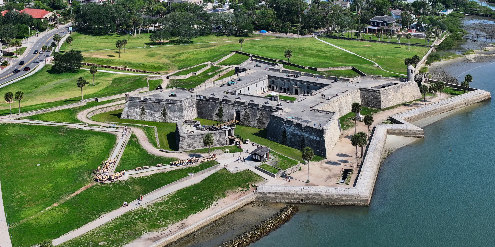 Vista aérea diurna do Castillo de San Marcos, com muralhas de pedra, gramados, palmeiras e a margem da baía em St. Augustine.
