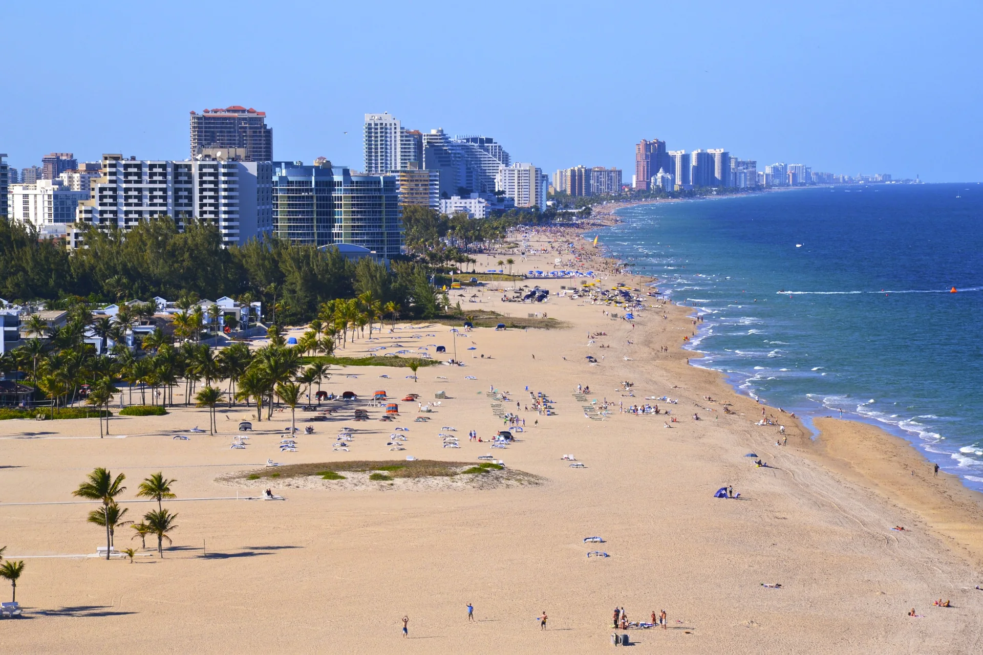Vista ampla de Fort Lauderdale Beach com areia clara, palmeiras, mar azul e a linha de edifícios da orla sob céu limpo.