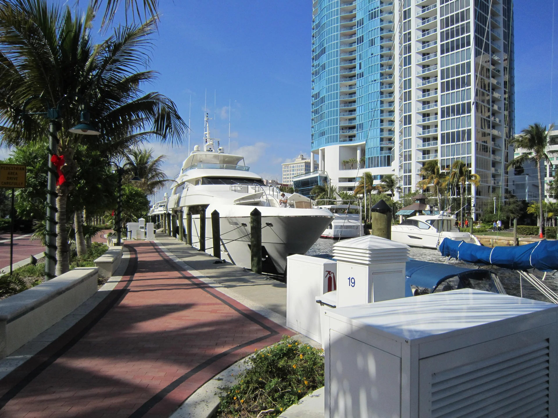 Trecho de passarela do Riverwalk de Fort Lauderdale junto ao New River, com palmeiras, bancos, uma embarcação e torres ao fundo.