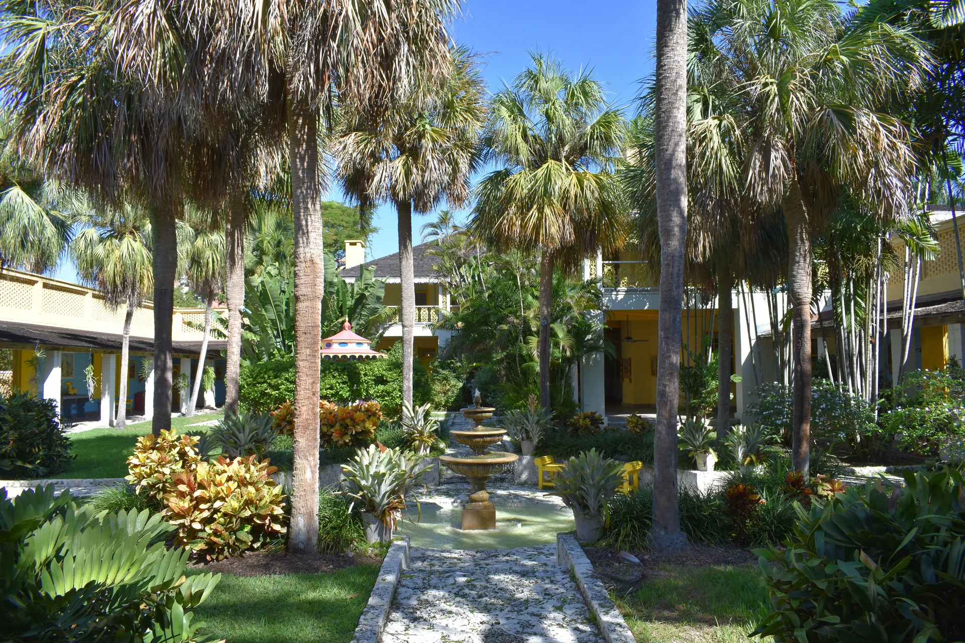 Pátio ajardinado da Bonnet House em Fort Lauderdale, com palmeiras, fonte central, vegetação tropical e edifícios amarelos sob sol.