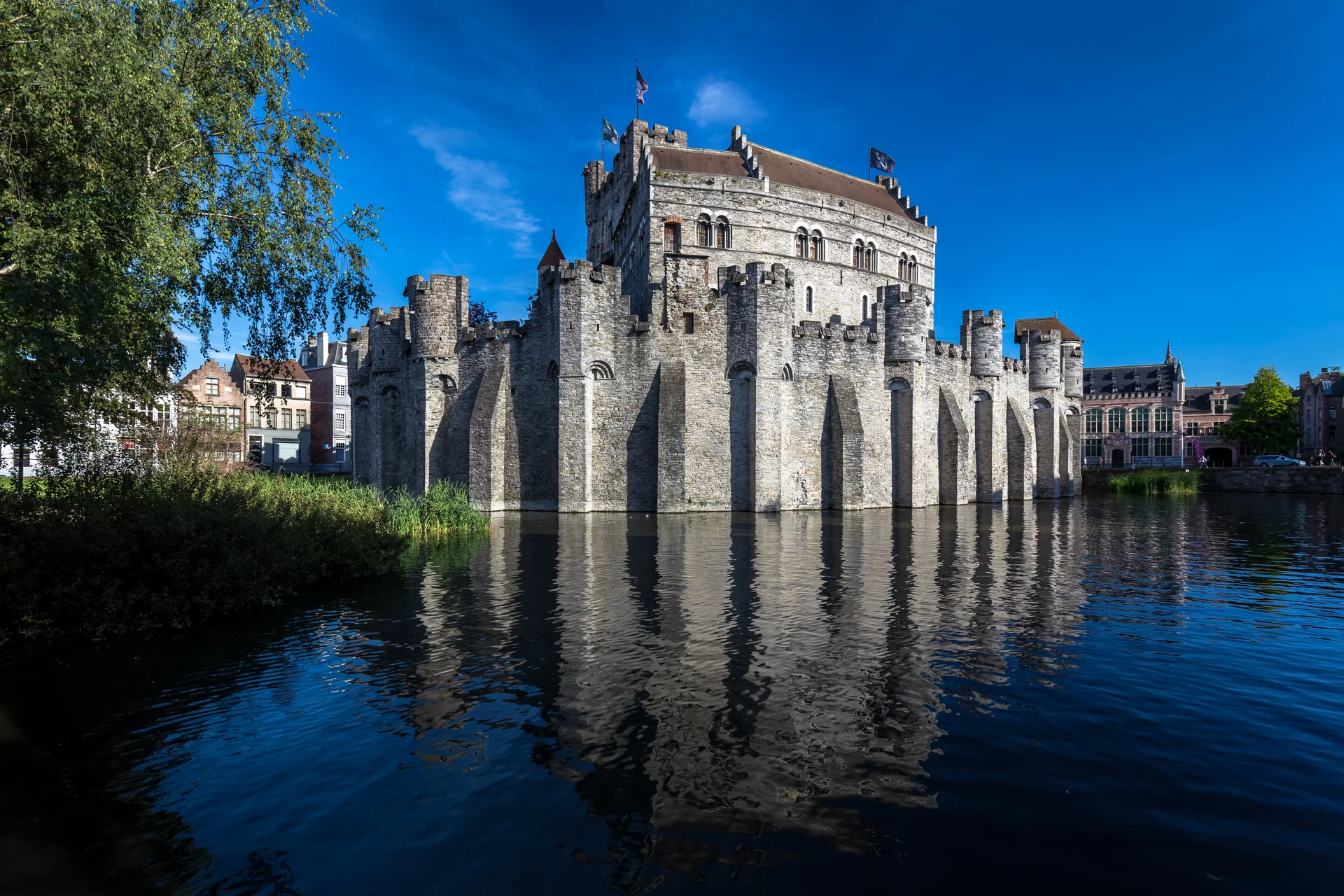 Gravensteen visto da Sint-Widostraat, com as muralhas de pedra refletidas na água sob céu azul.