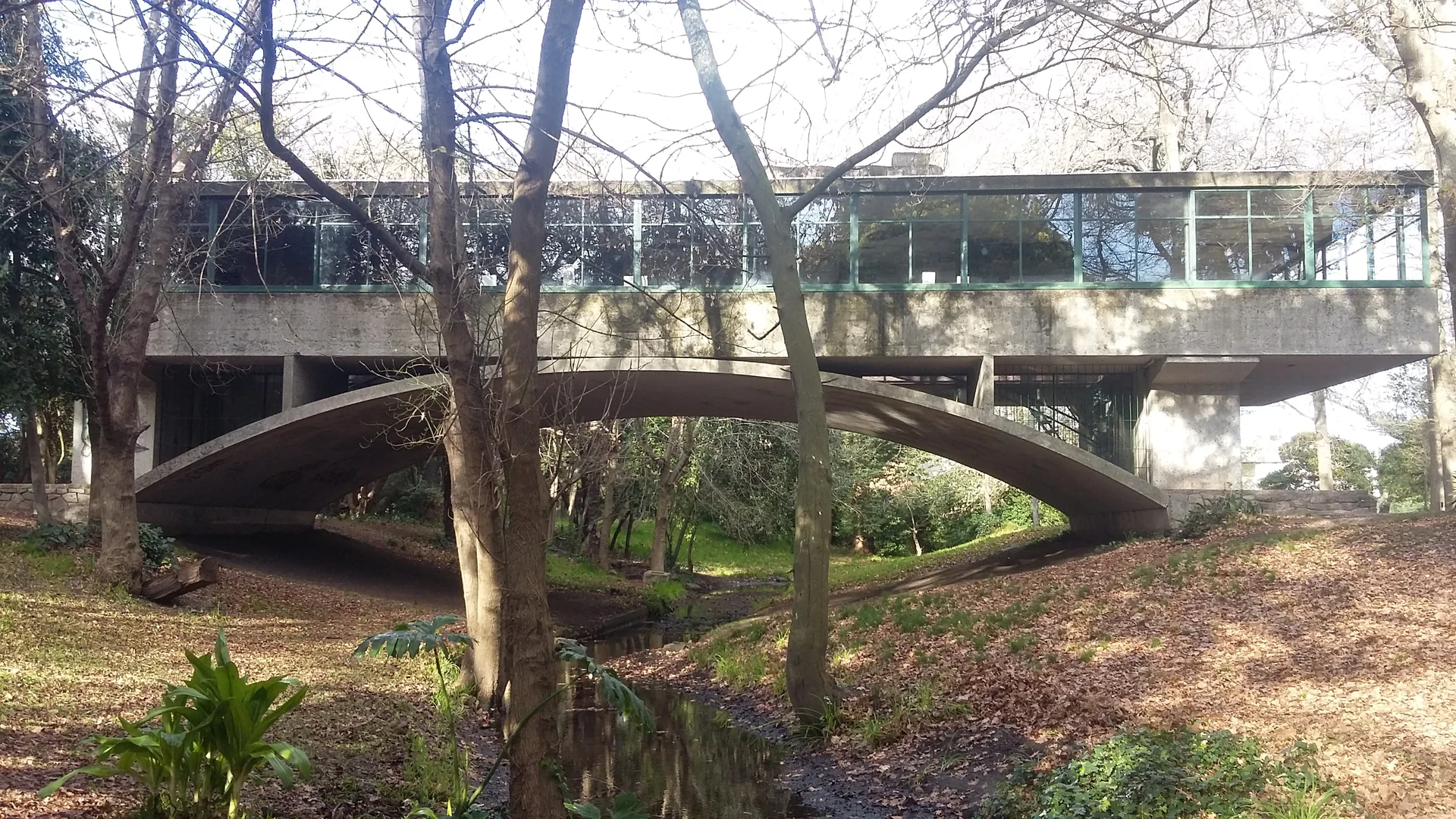 Casa sobre el Arroyo em Mar del Plata, com o volume envidraçado apoiado sobre o arco de concreto e árvores ao redor.