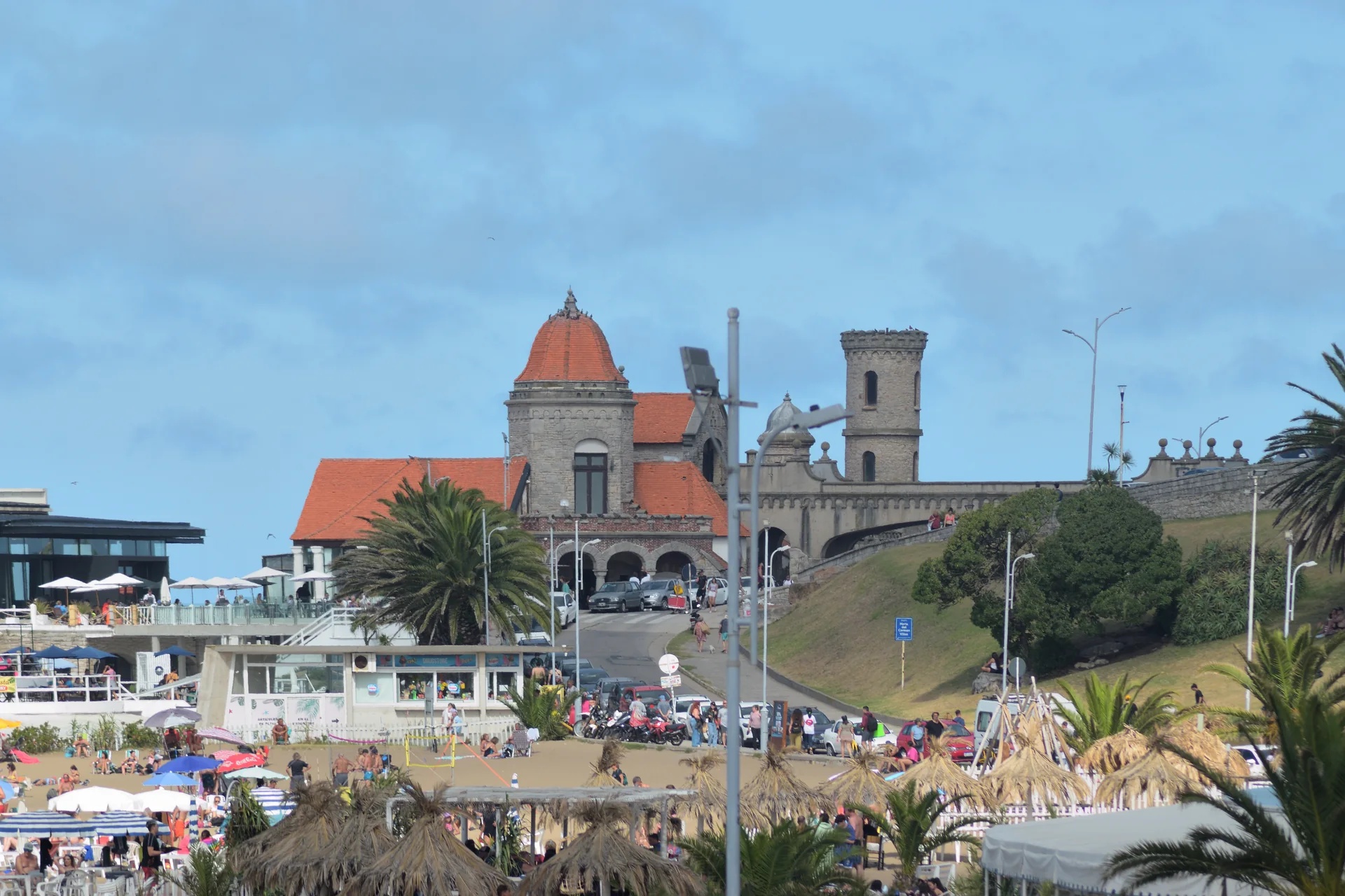 Torreón del Monje em Mar del Plata, com torres de pedra, telhados alaranjados, palmeiras e praia no primeiro plano.