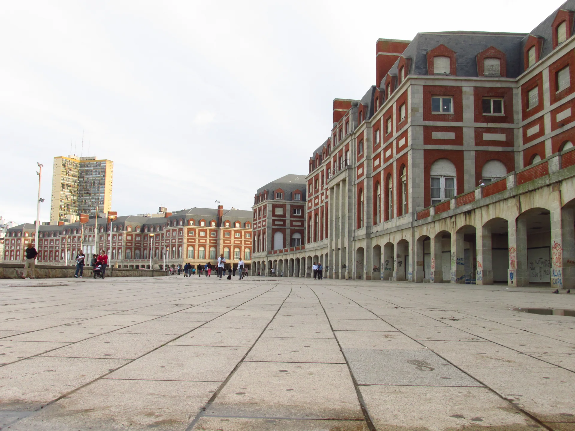 Calçadão da Rambla de Mar del Plata junto aos edifícios de tijolos vermelhos e arcos, com pedestres no espaço público.