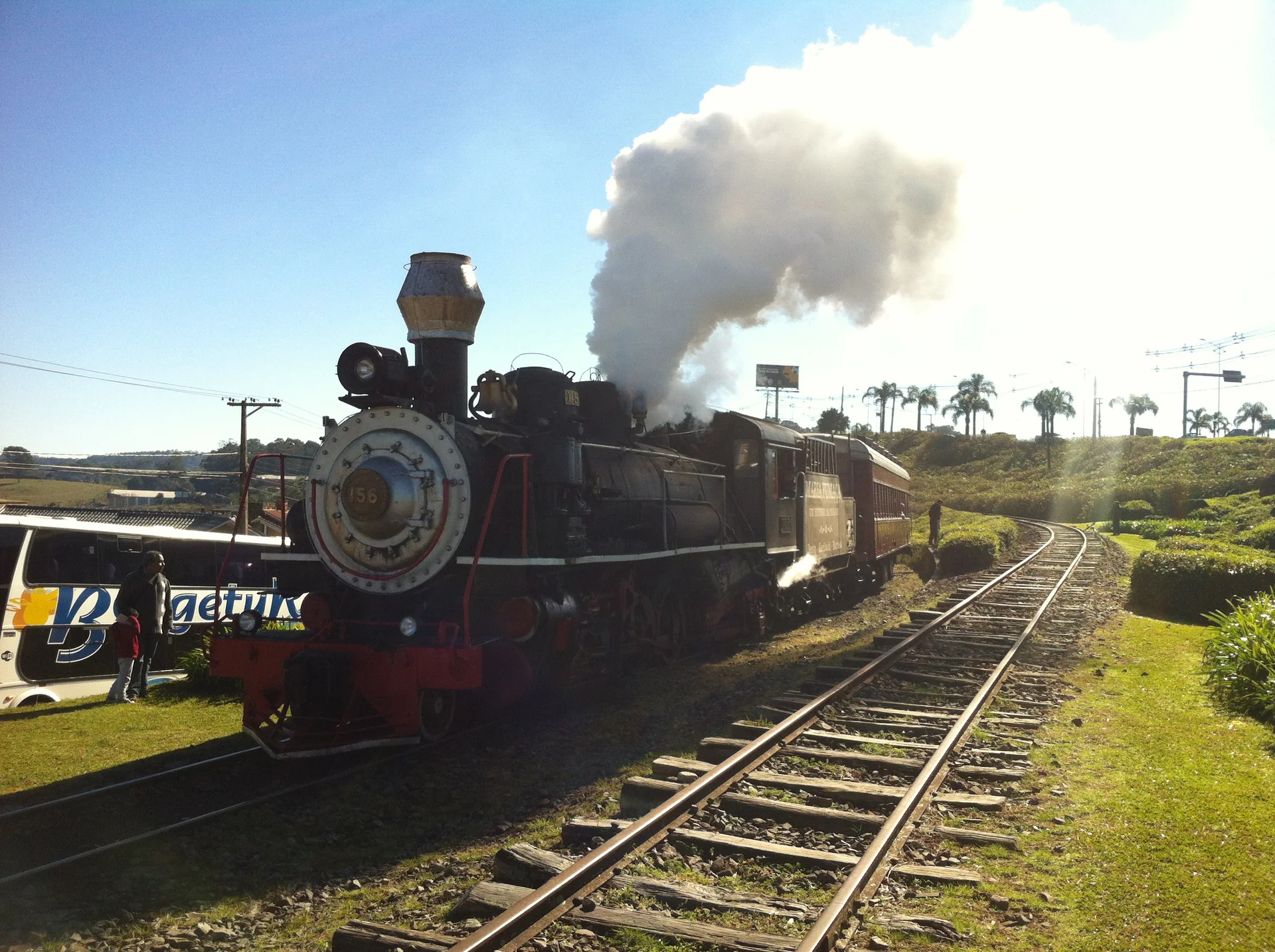 Maria Fumaça do Trem do Vinho em Carlos Barbosa, com vapor saindo da locomotiva e trilhos em curva sob luz do dia.