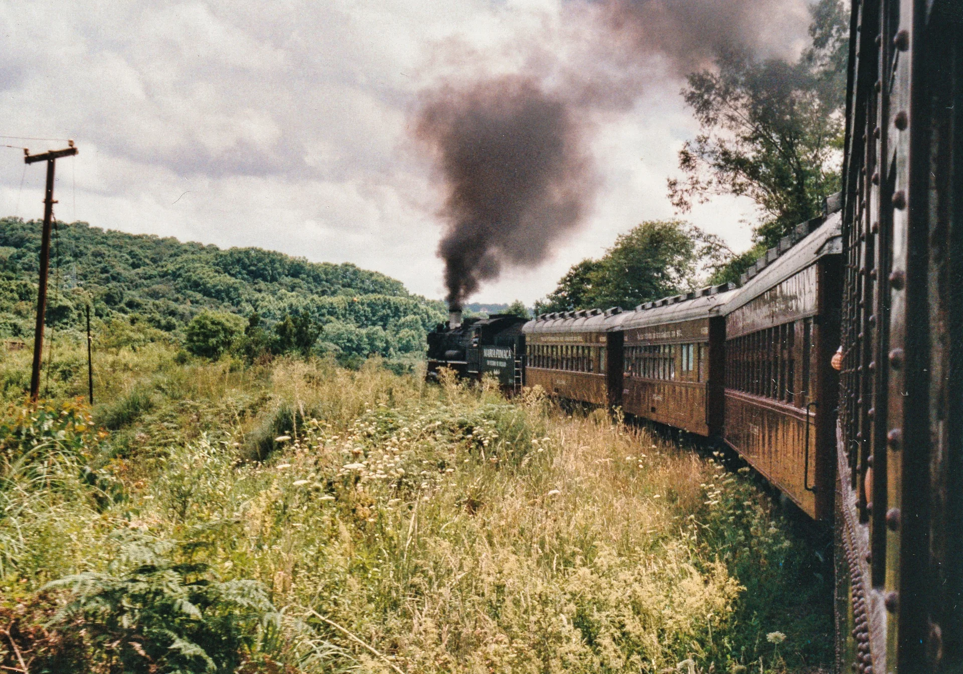 Vagões da Maria Fumaça avançam por área verde rumo a Carlos Barbosa, vistos de uma janela do trem, com fumaça da locomotiva ao fundo.