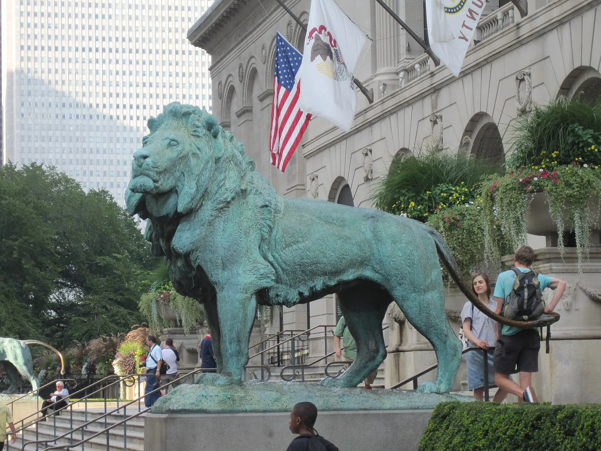 Leão de bronze diante da fachada do Art Institute of Chicago, com bandeiras e jardins floridos em cena diurna.