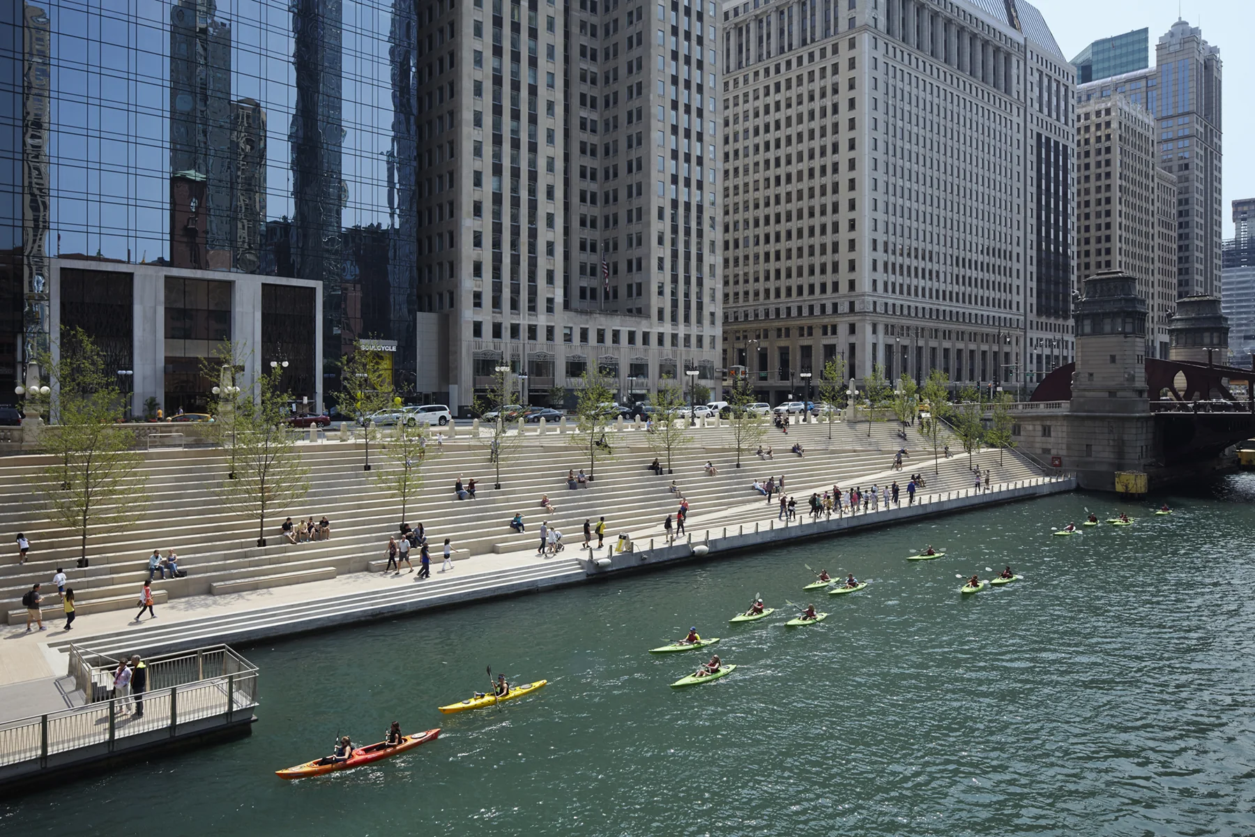 Chicago Riverwalk em tarde clara, com caiaques no rio, pessoas nas escadarias e edifícios do centro ao fundo.