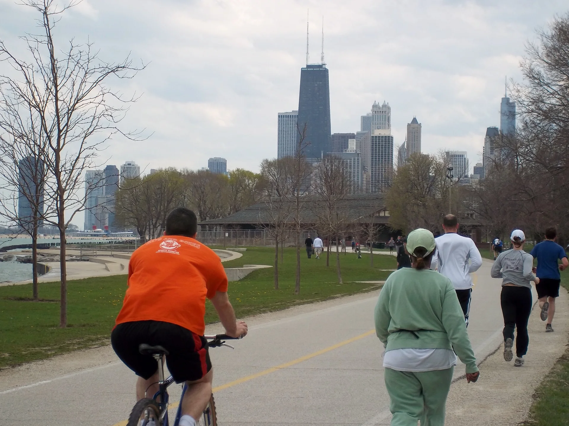 Ciclista e pessoas caminhando na Lakefront Trail perto de Lincoln Park, com gramado, lago e skyline de Chicago ao fundo.
