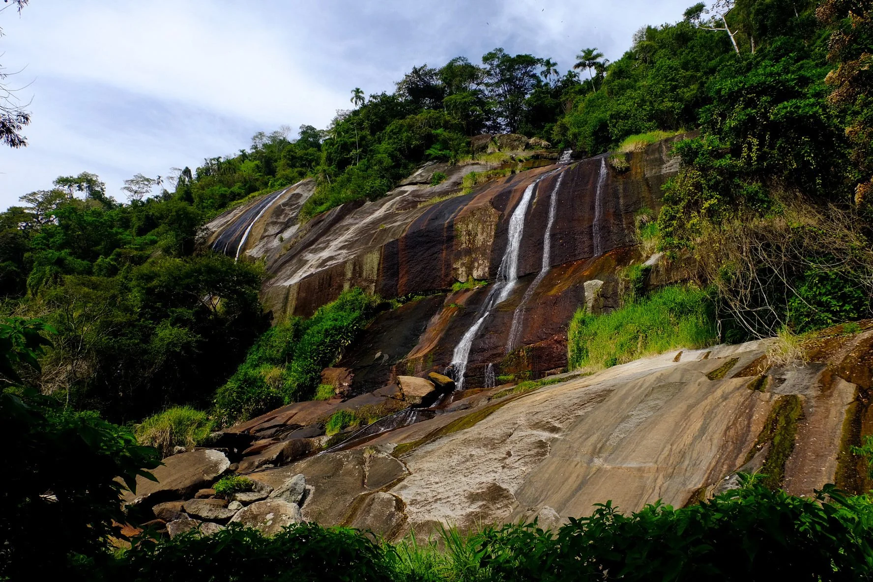 Cachoeira da Água Branca cai por uma parede de rocha escura cercada por vegetação verde no Parque da Usina, em Ilhabela.