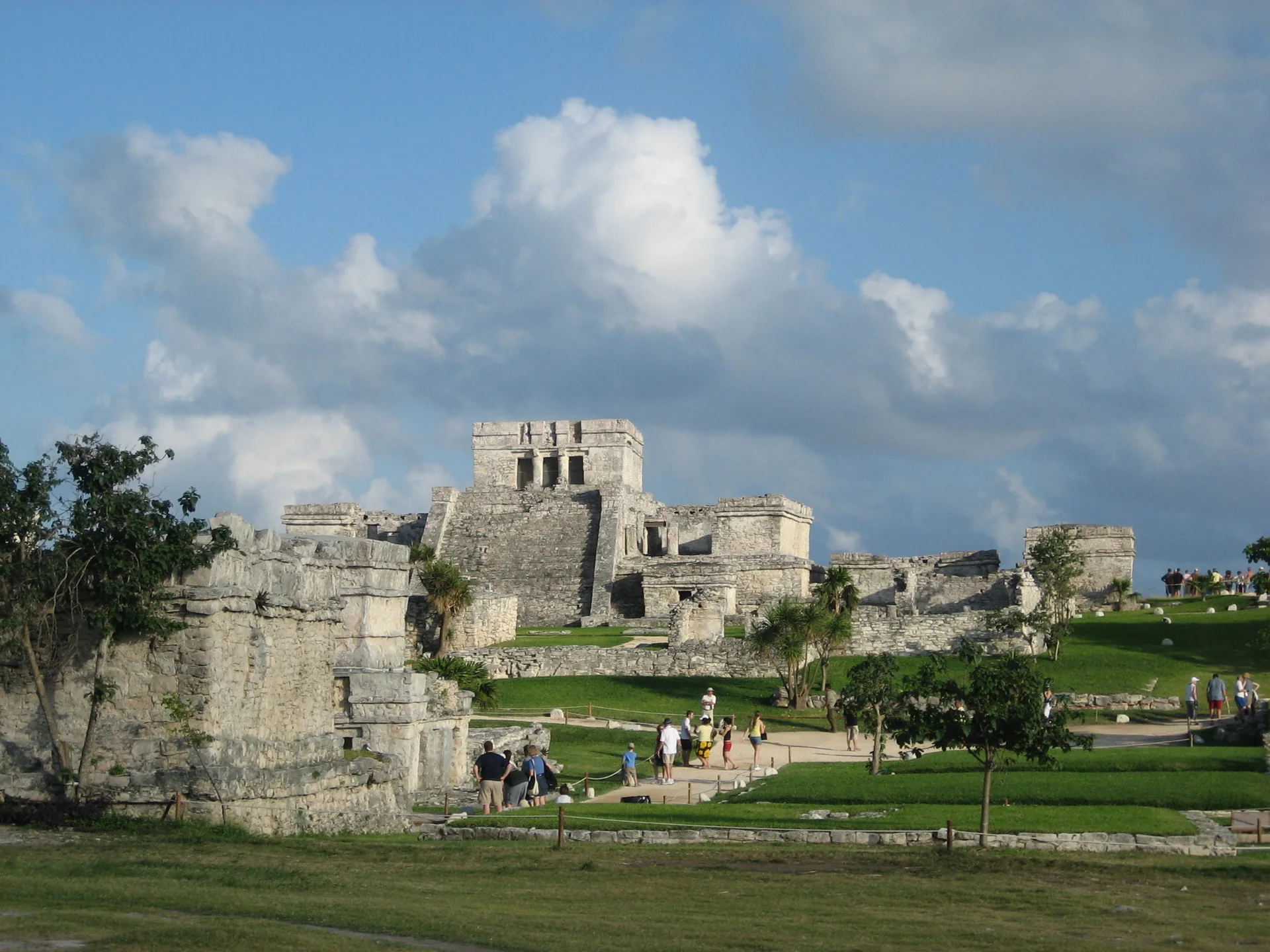 Estruturas maias de pedra de Tulum, com El Castillo ao centro, gramado e visitantes no percurso delimitado sob luz diurna.