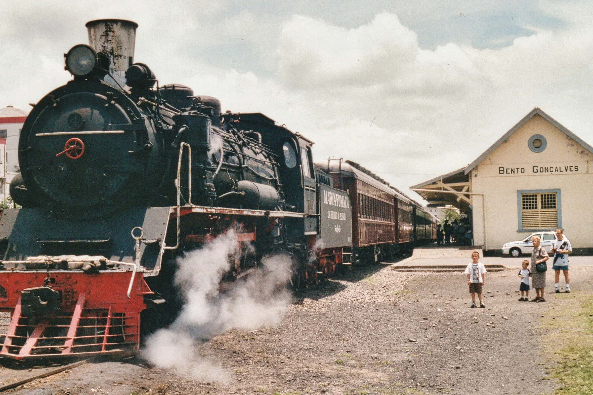 Locomotiva Maria Fumaça junto à estação de Bento Gonçalves, com vapor e passageiros no pátio.