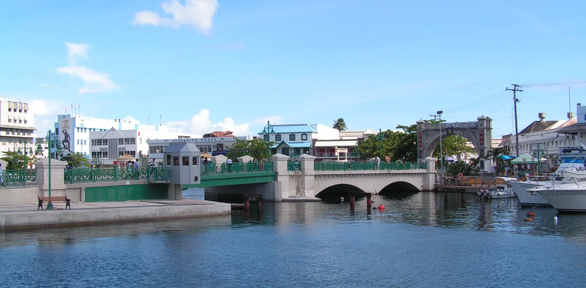 Vista do Careenage em Bridgetown com água, embarcações, a Chamberlain Bridge e a Independence Arch sob céu azul.