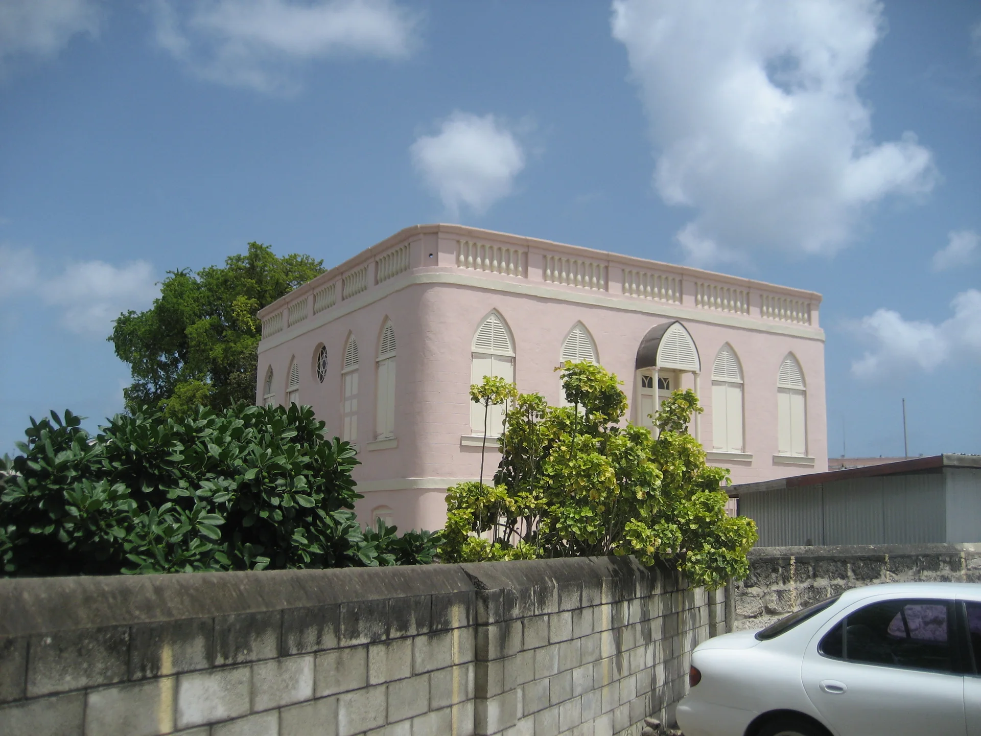 Vista externa da Nidhe Israel Synagogue em Bridgetown, edifício rosa com janelas em arco, vegetação e céu azul.