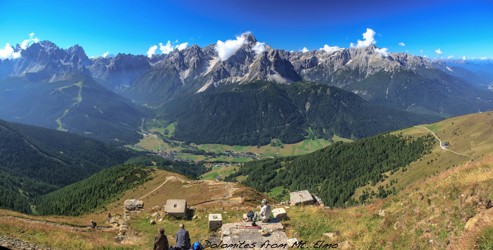 Vista ampla das Dolomitas a partir do Monte Elmo, com vale alpino, campos e cadeias rochosas ao fundo.