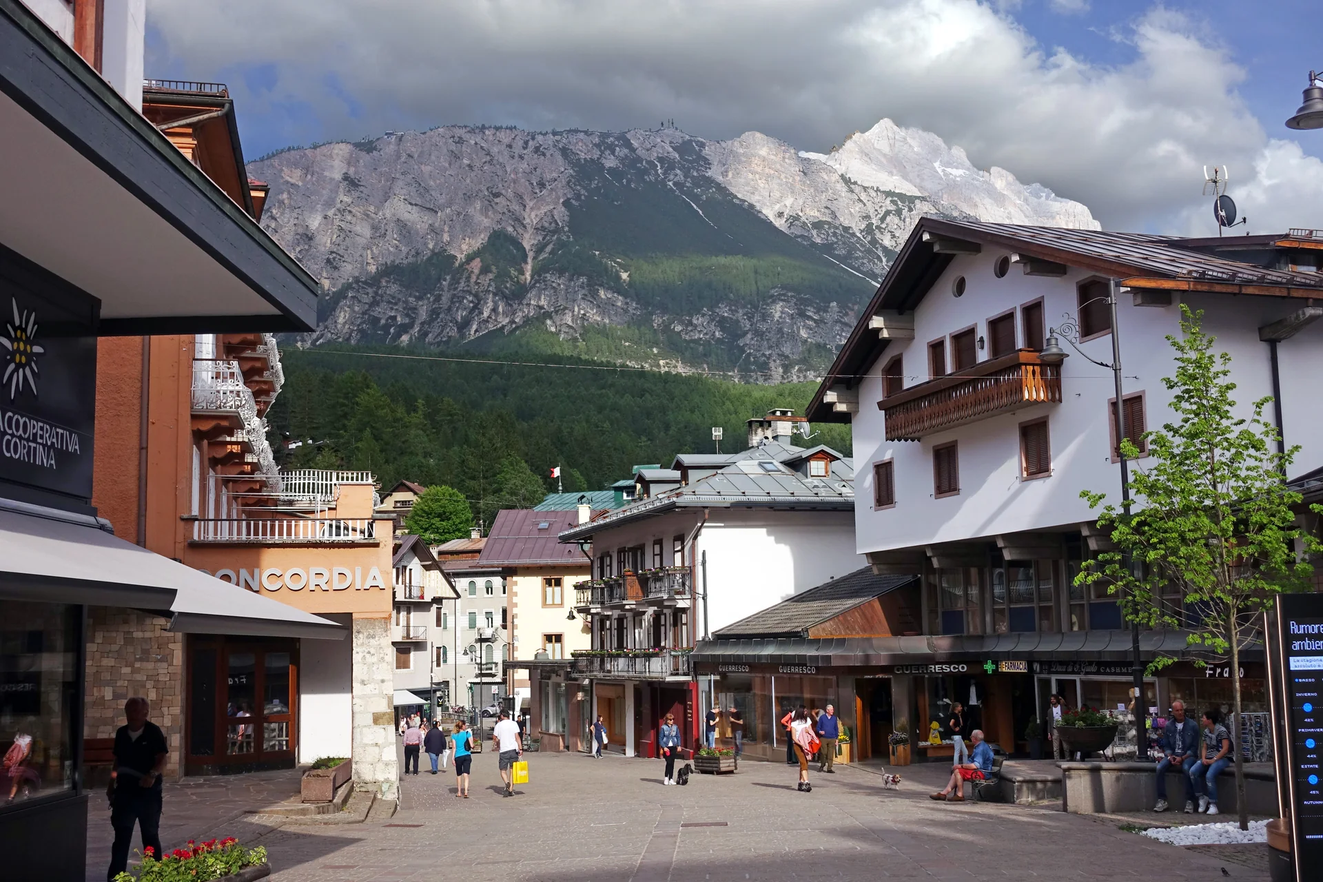Rua Corso Italia em Cortina d’Ampezzo, com edifícios da vila, pedestres e montanhas ao fundo em luz de fim de tarde.