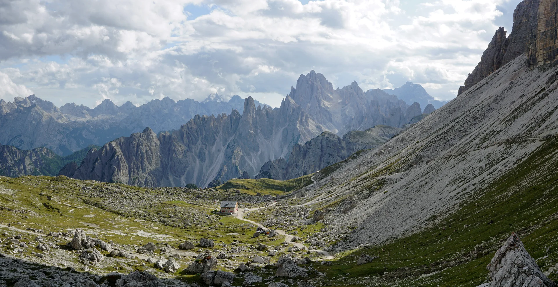 Rifugio Lavaredo em contexto de rota alpina, com trilha de montanha, prado e o grupo Cadini di Misurina ao fundo.