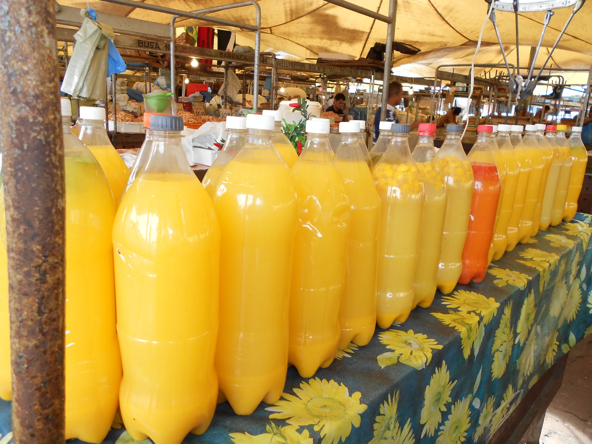 Fileira de garrafas de tucupi amarelo em uma banca de mercado, com o ambiente da feira ao fundo.