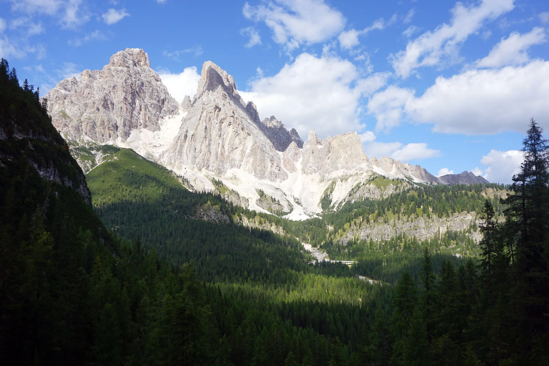 Paredões claros do Monte Cristallo vistos da rota de caminhada do Lago di Sorapis, acima de um vale arborizado sob céu azul.