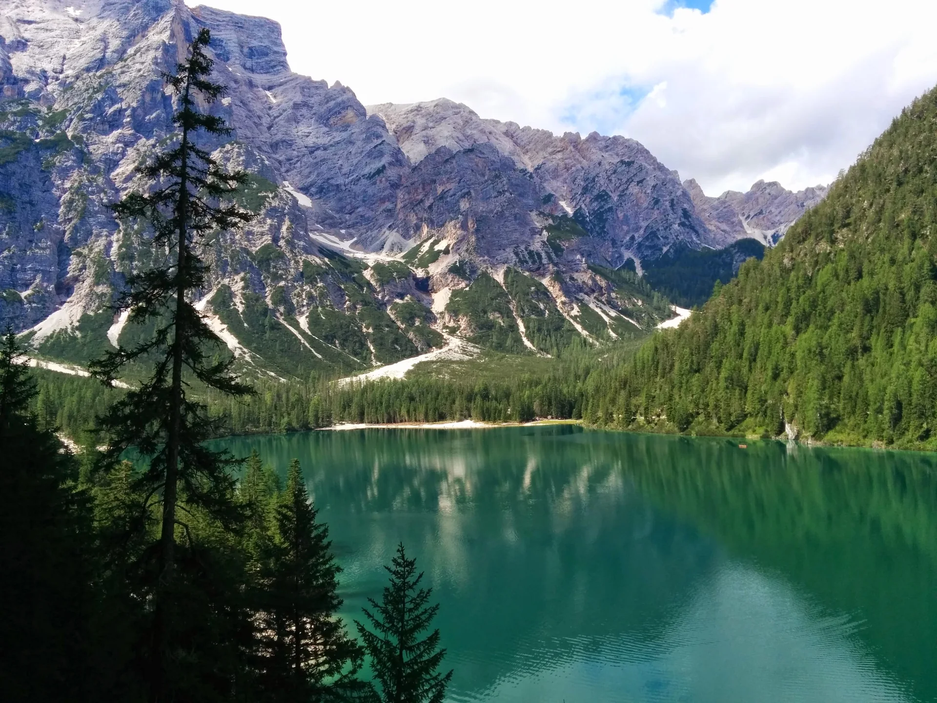 Vista do Lago di Braies com água verde, coníferas na margem e montanhas rochosas sob céu parcialmente nublado.