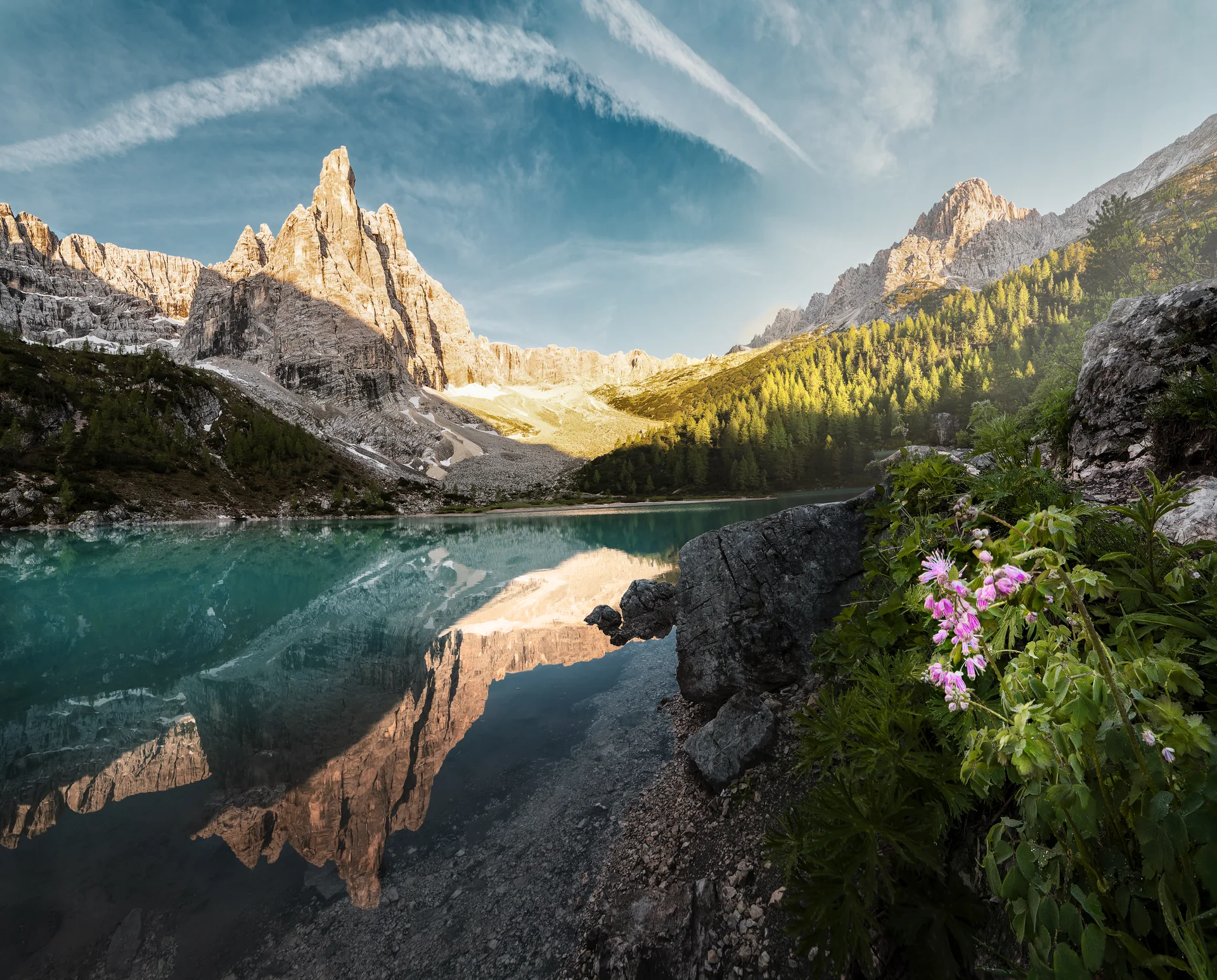 Lago di Sorapis refletindo os paredões rochosos, com flores alpinas na margem e luz suave de início da manhã.