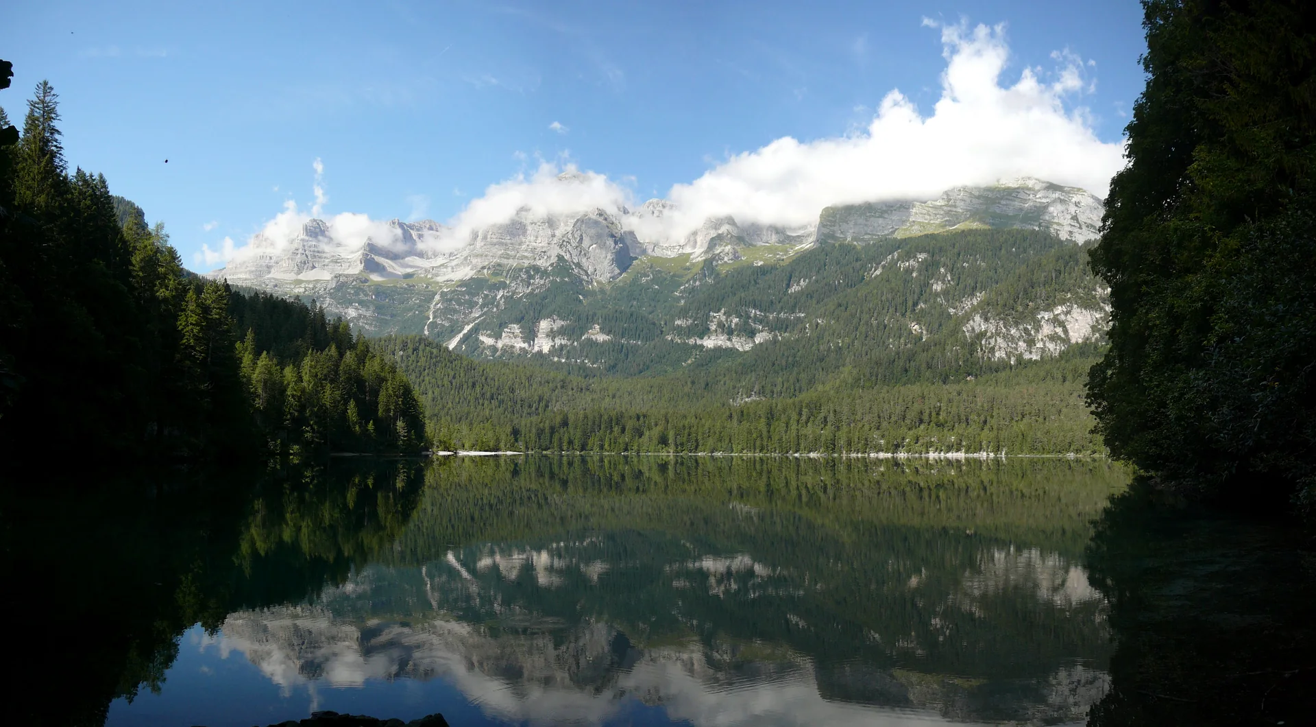 Lago di Tovel visto da margem leste, com reflexo de floresta e montanhas parcialmente envoltas por nuvens em dia claro.
