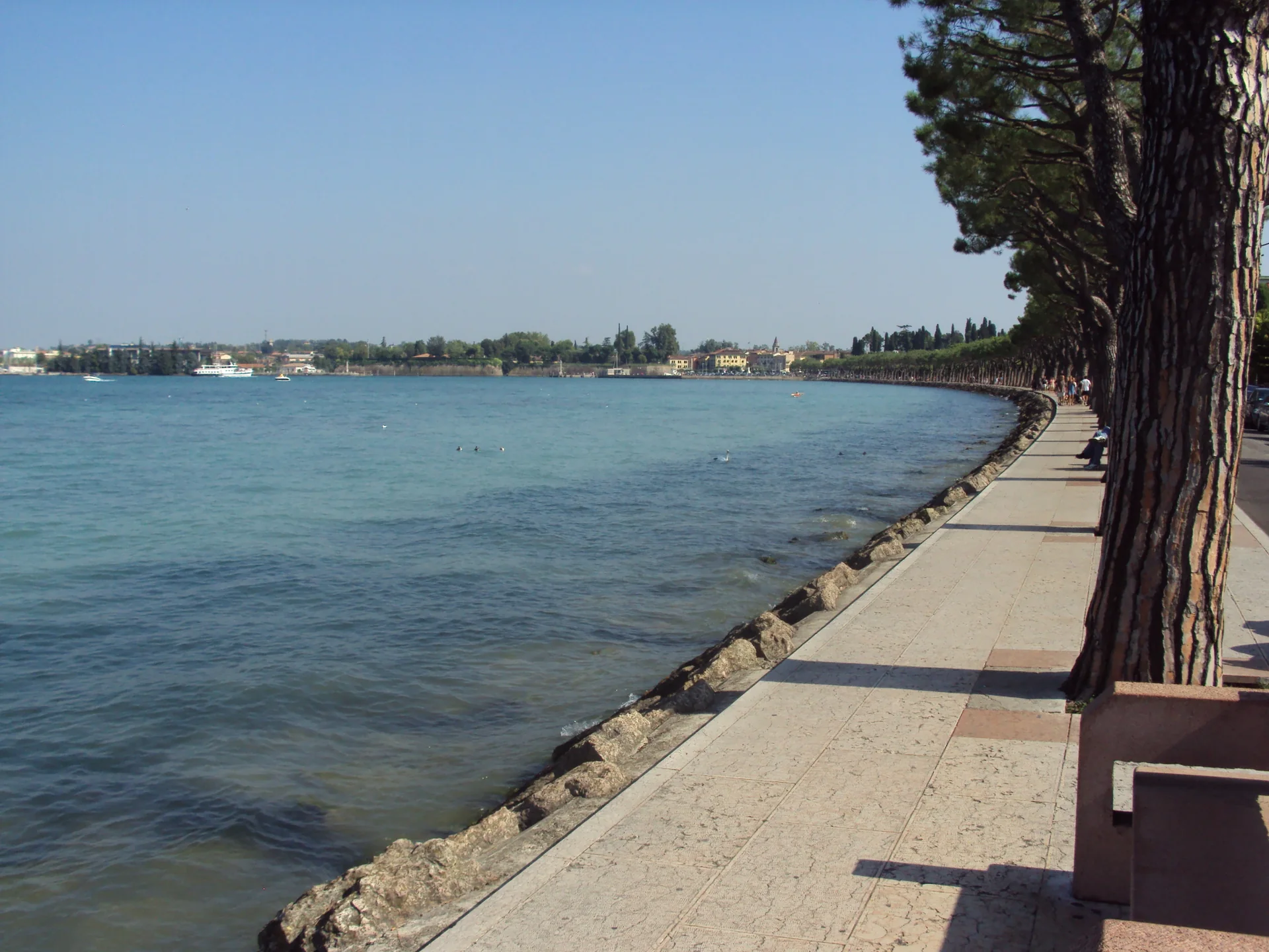 Calçadão arborizado junto ao Lago di Garda em Peschiera, com água, margem distante e pedestres ao fundo.