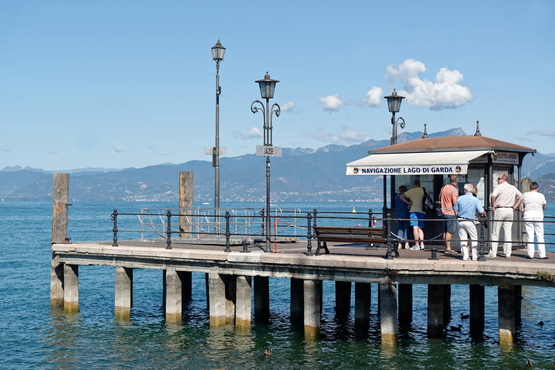 Cais da Navigazione Lago di Garda em Lazise, com cabine de embarque, passageiros, água azul e montanhas ao fundo.