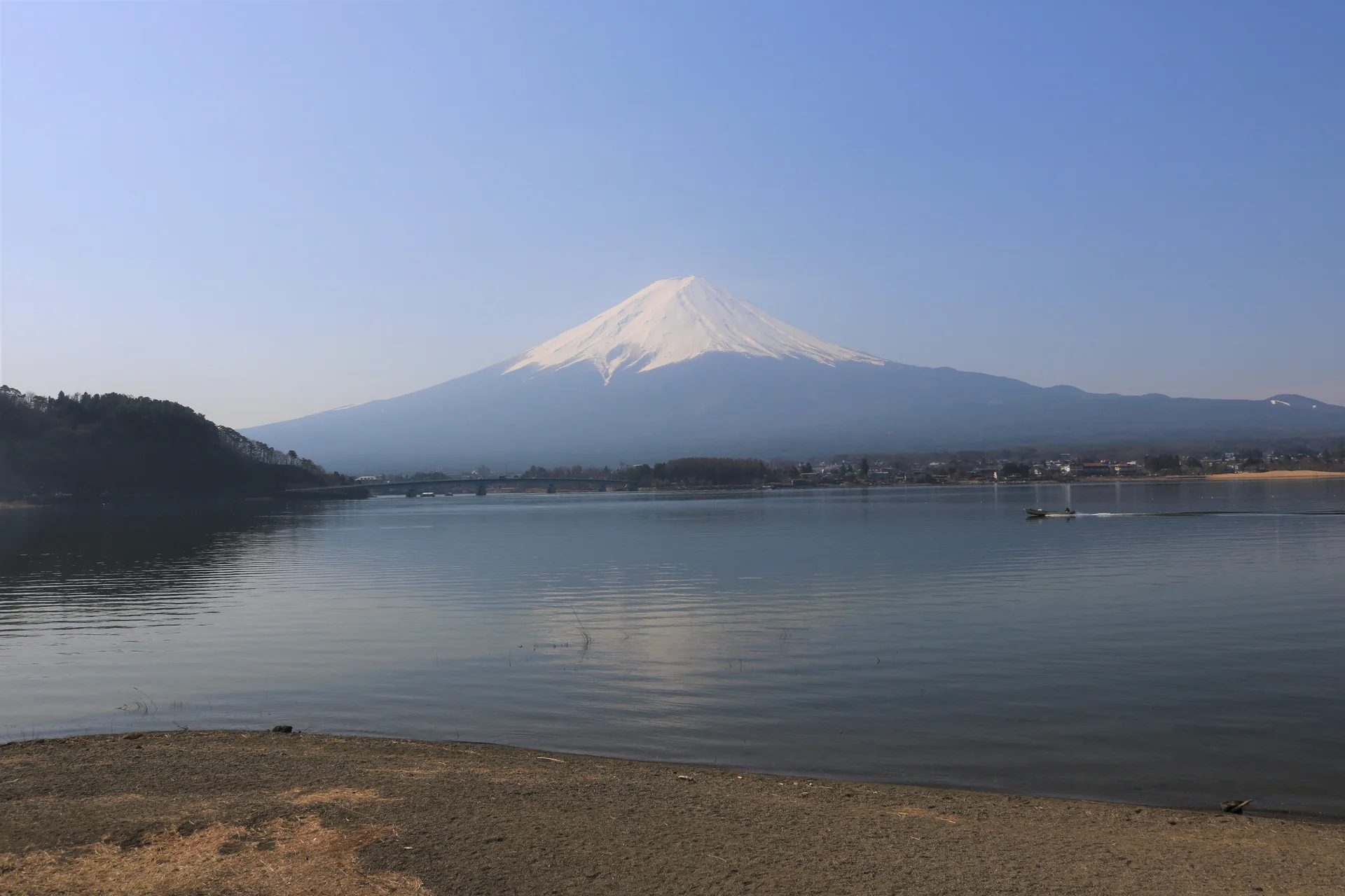 Monte Fuji visto da margem do Lago Kawaguchi, com água calma, ponte e céu azul-claro.