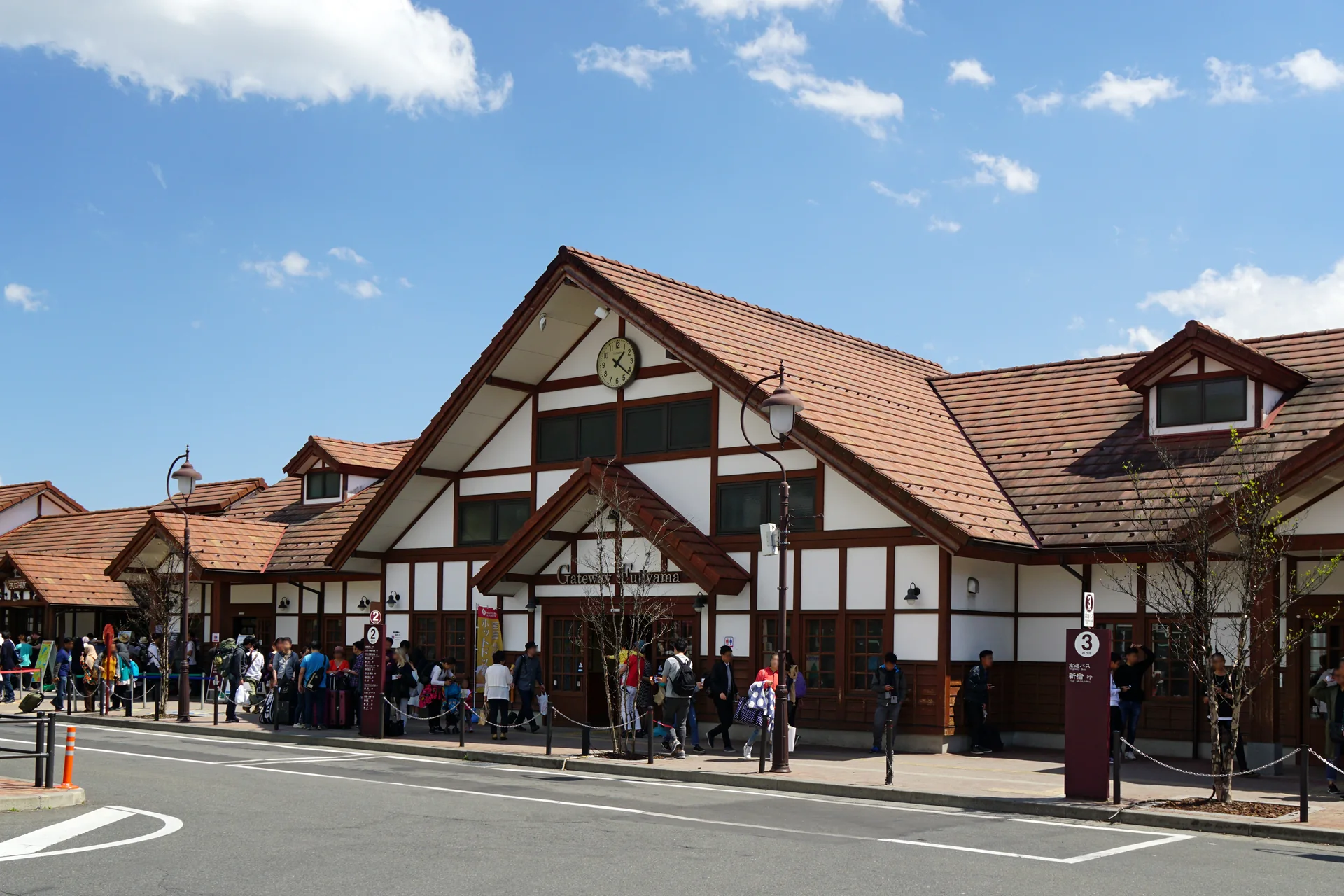 Fachada da Estação Kawaguchiko com relógio, telhado de madeira e passageiros em plena luz do dia.