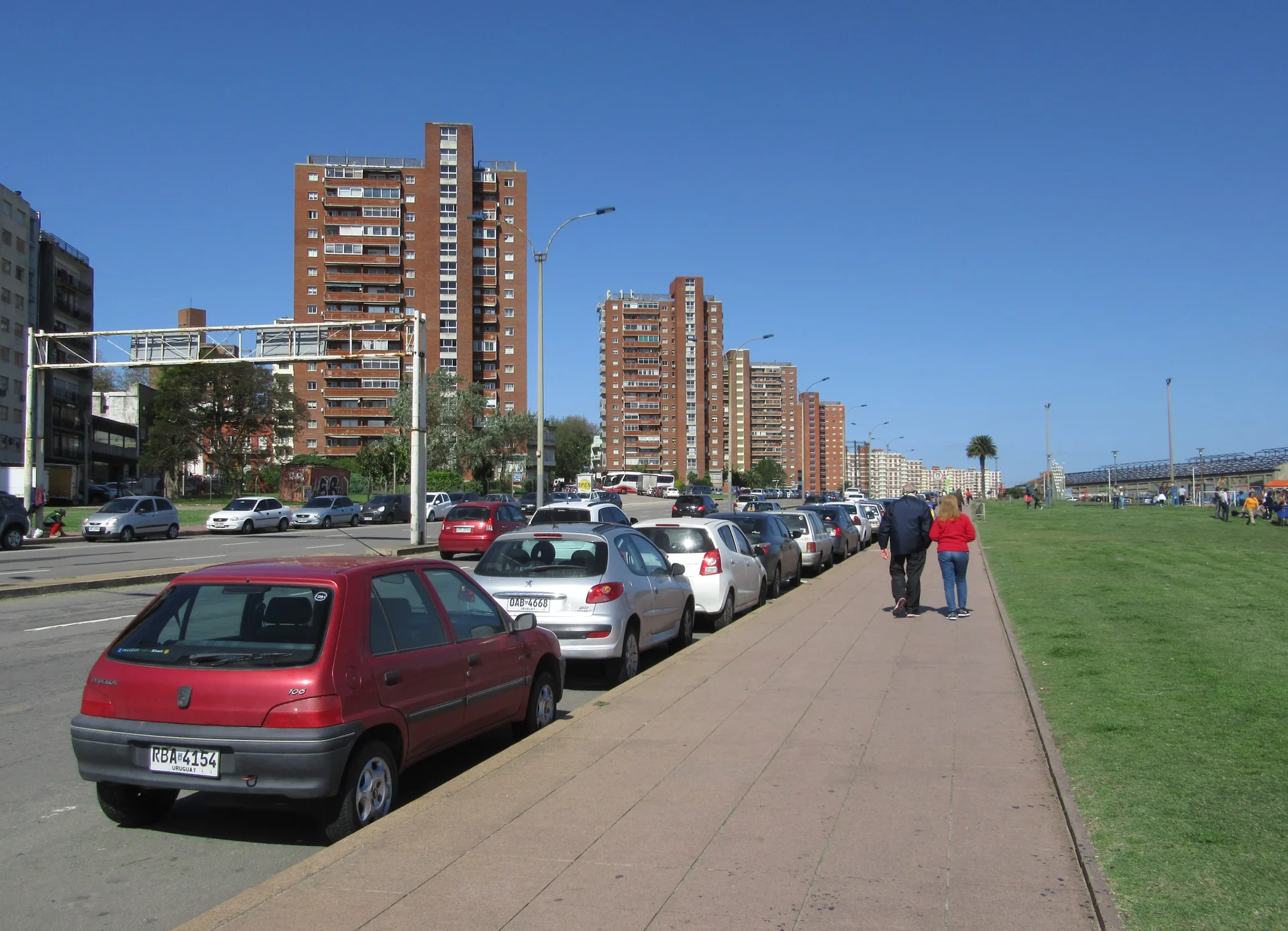Calçada da rambla Gran Bretaña em Montevidéu, com pessoas caminhando, gramado, avenida e edifícios residenciais sob céu azul.