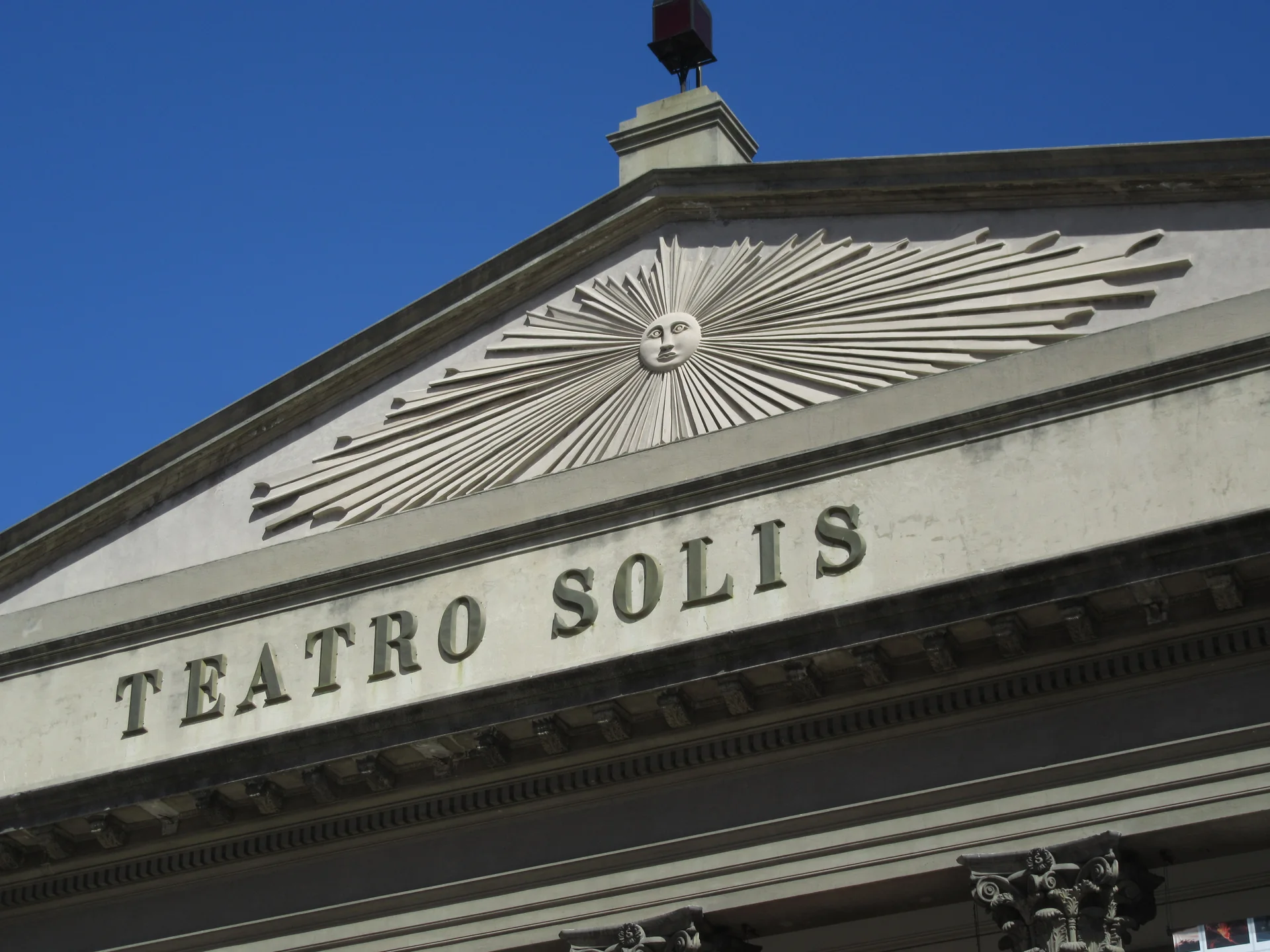 Frontão da fachada do Teatro Solís de Montevidéu, com o nome do teatro, relevo solar e céu azul em luz diurna.