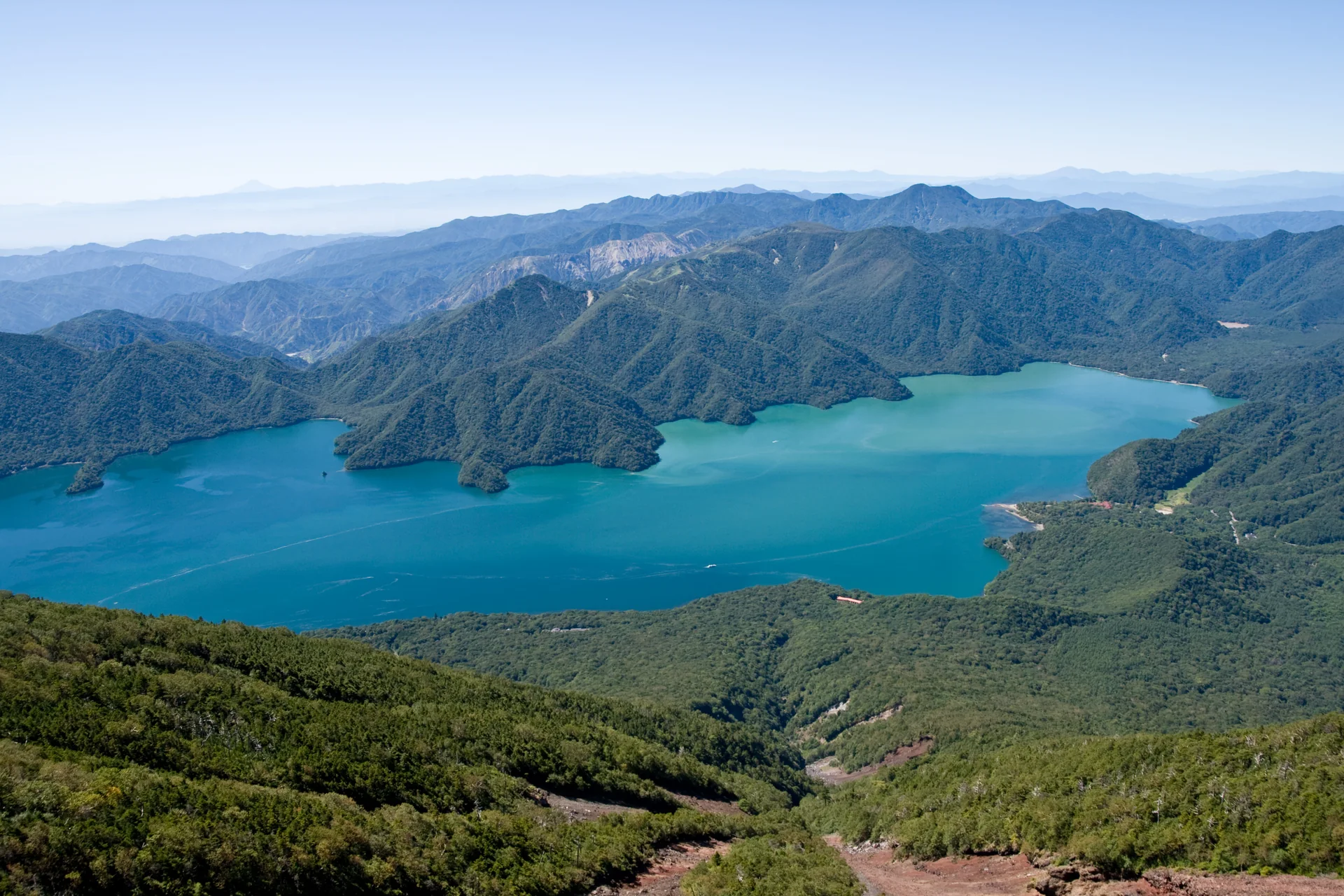 Vista elevada do Lago Chuzenji em Oku-Nikko, com água azul e montanhas sob céu claro.