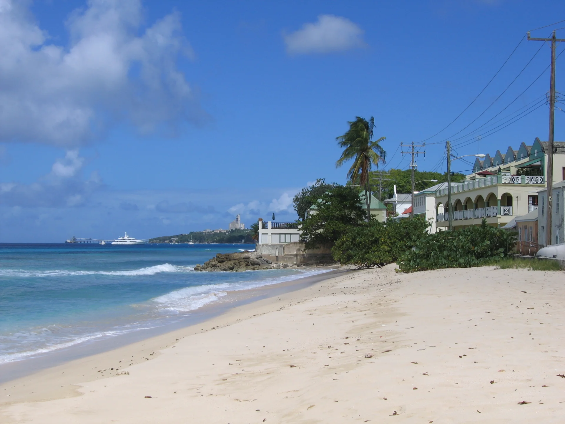 Praia de Speightstown, em Saint Peter, com faixa de areia, água azul, casas costeiras e fiação ao fundo.
