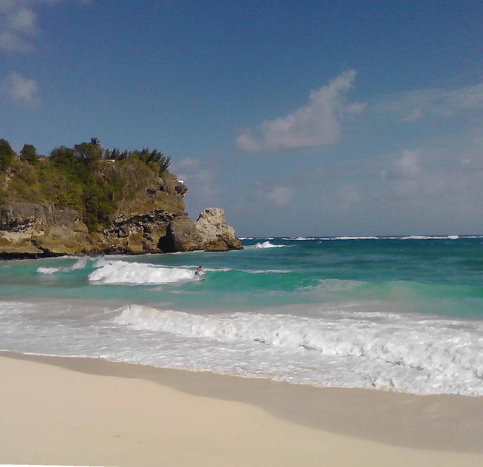 Praticante de surfe em Foul Bay, Barbados, em ondas diante de costão rochoso, visto a distância da areia.