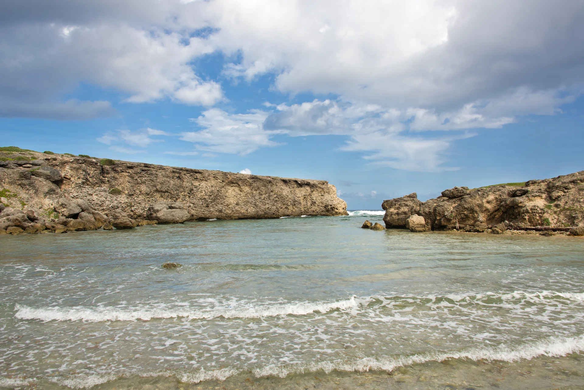 Enseada de River Bay, em St. Lucy, com água rasa, rochas calcárias e ondas visíveis além da abertura para o mar.