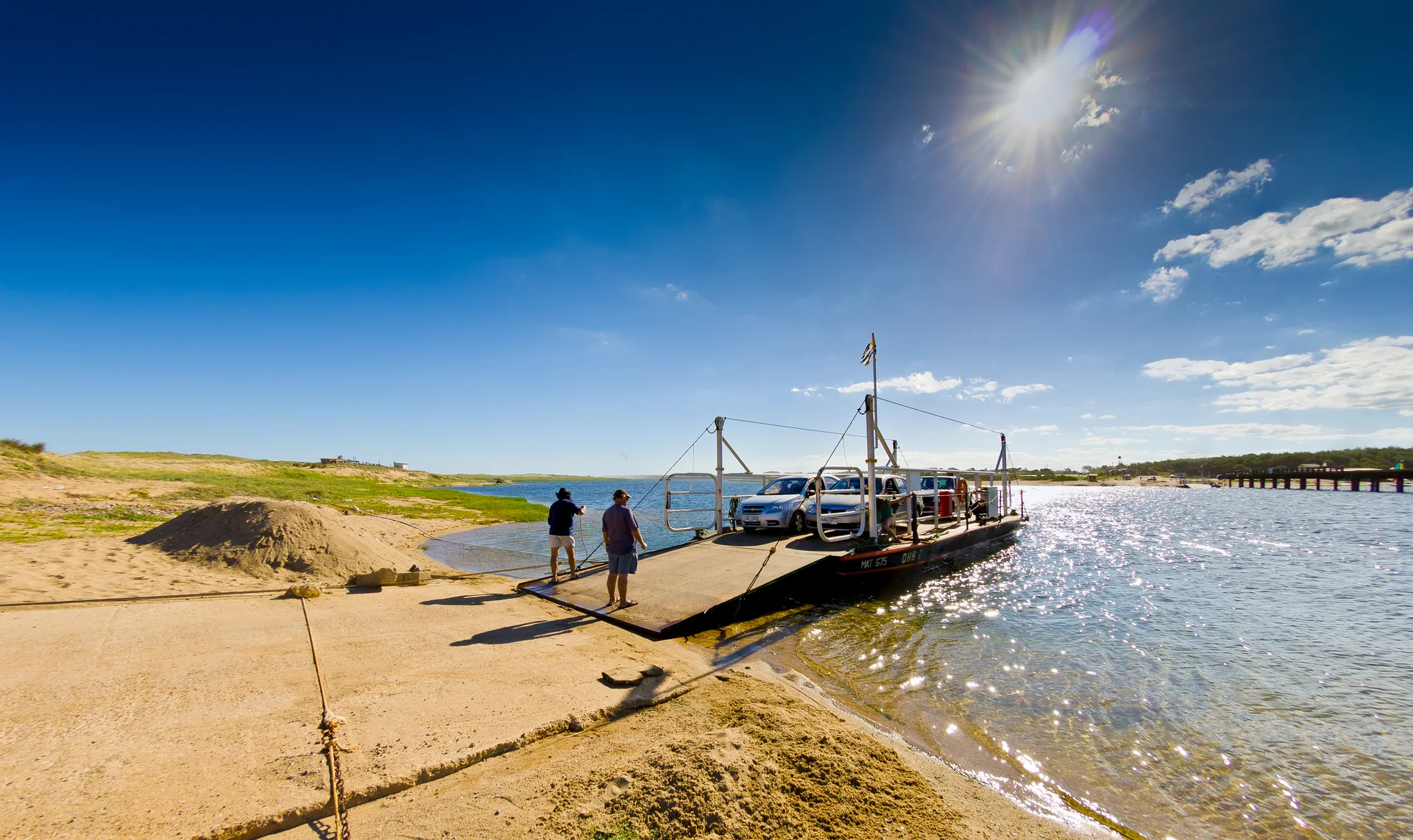 Margem arenosa e água da Laguna Garzón sob sol forte, com uma balsa junto à desembocadura e dunas ao fundo.