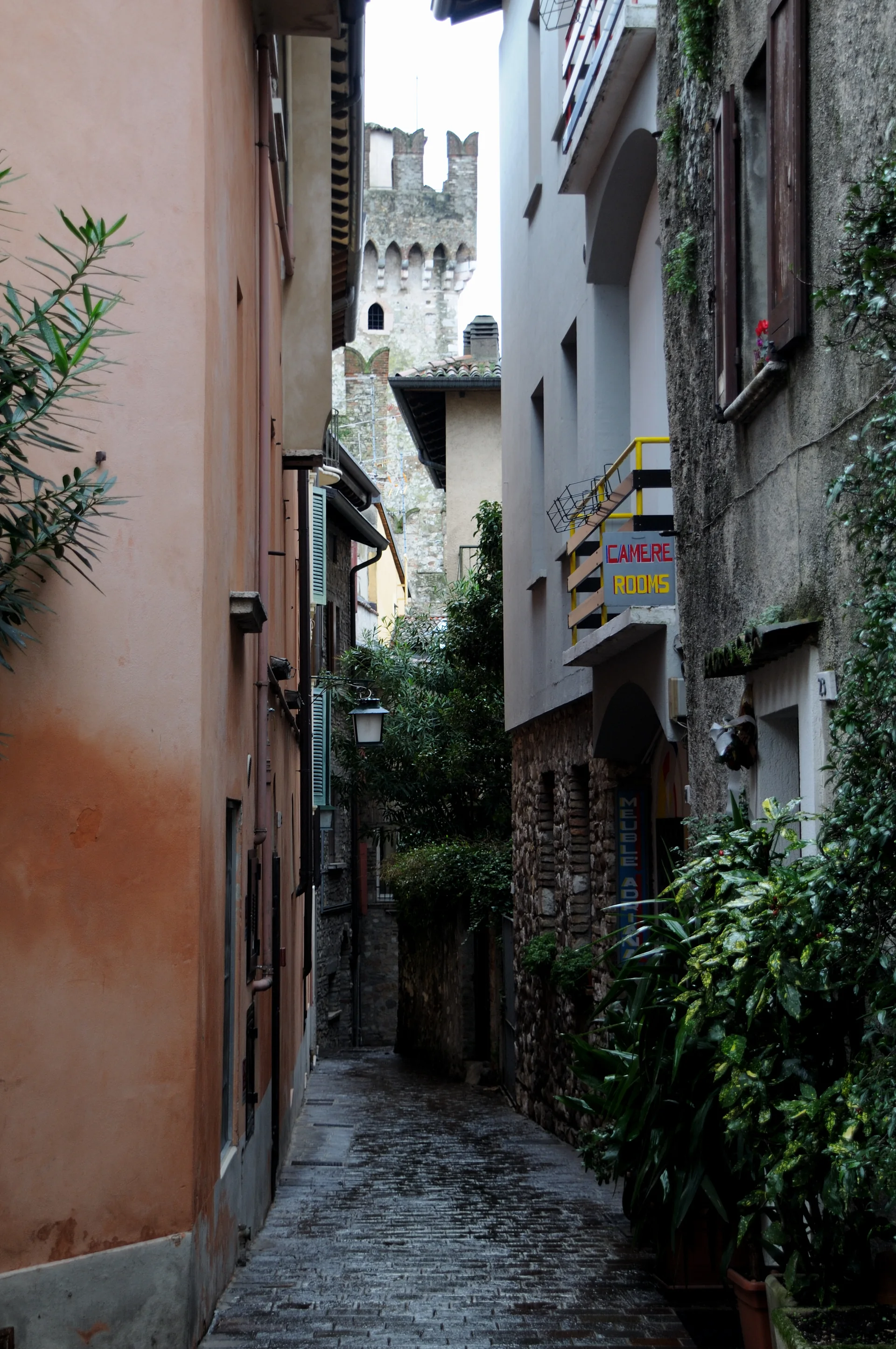 Viela de pedra sem pedestres em primeiro plano no centro histórico de Sirmione, com fachadas estreitas e torre do castelo ao fundo.