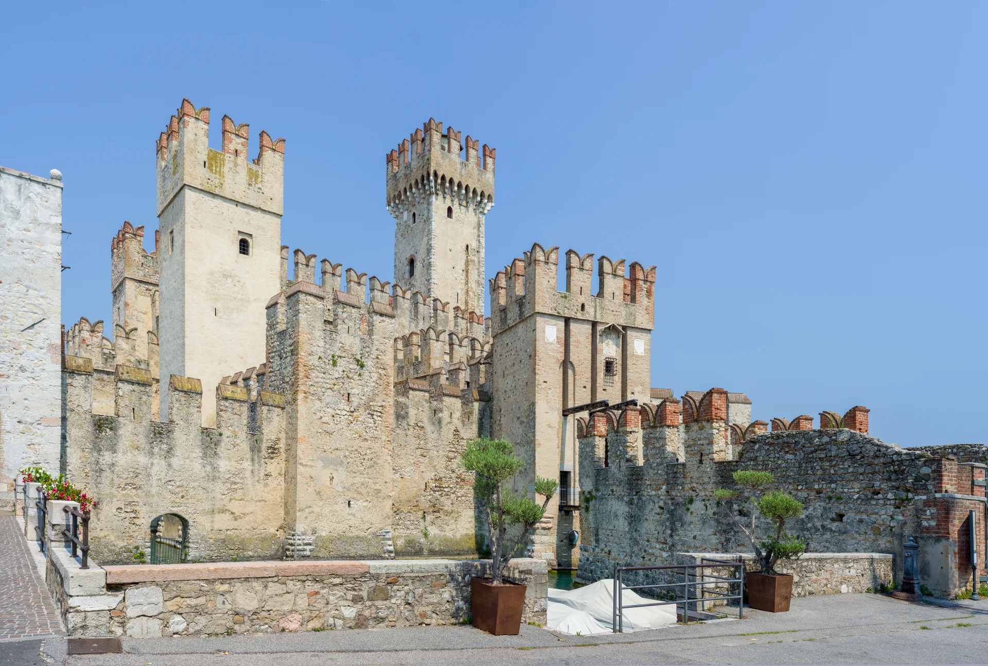 Torres e muralhas de pedra do Castello Scaligero de Sirmione, com trecho da dársena e céu azul em luz diurna.