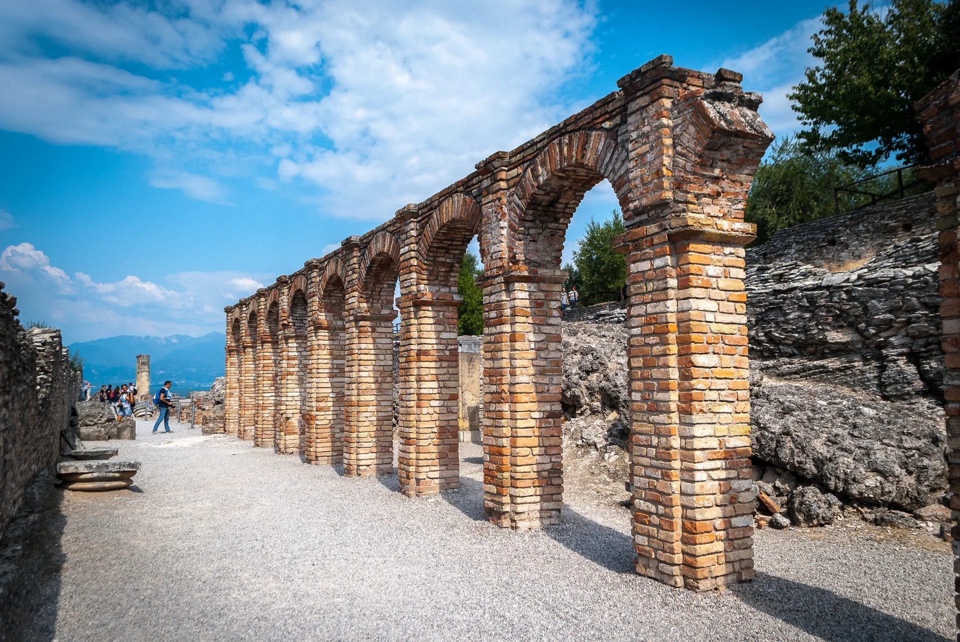 Ruínas romanas de tijolo das Grutas de Catullo em Sirmione, com caminho de visitação, oliveiras e o Lago di Garda ao fundo.