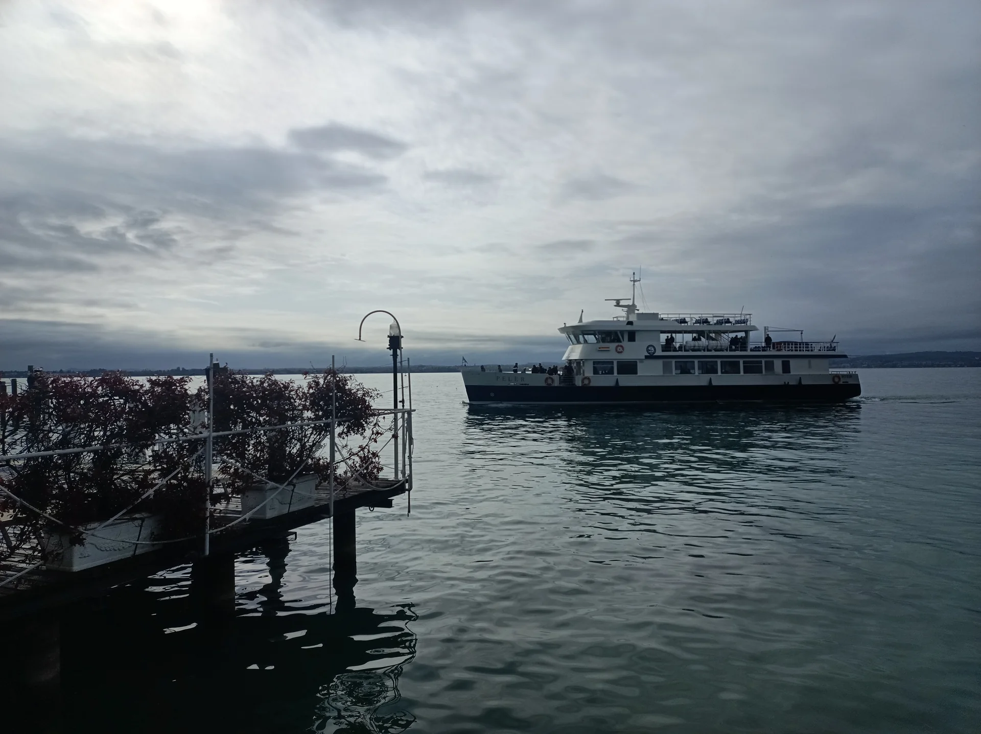 Embarcação regular navega junto ao cais público de Sirmione no Lago di Garda, em luz diurna sob céu encoberto claro.