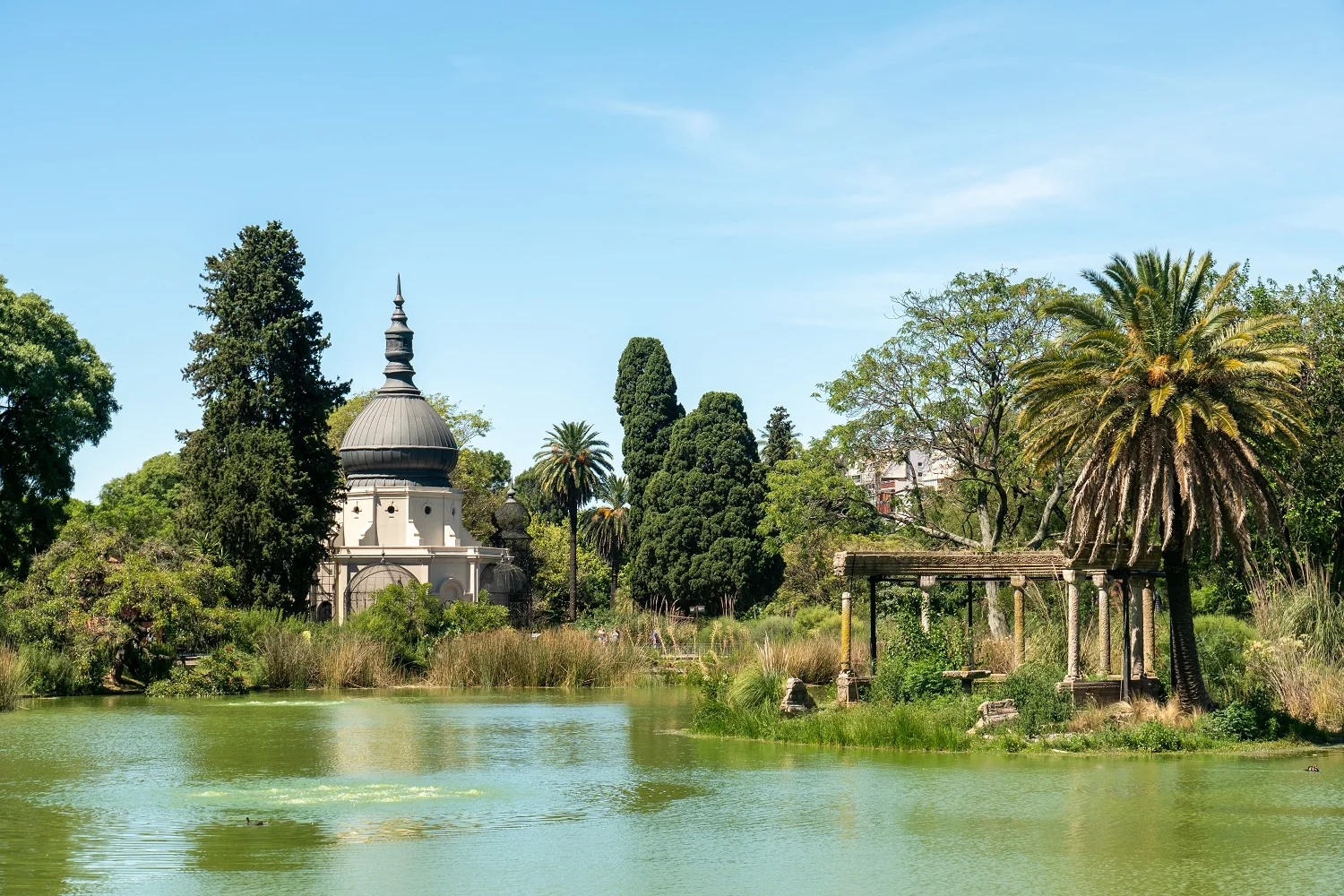 Pavilhão histórico e pórtico bizantino à beira do lago Darwin, entre vegetação do Ecoparque de Buenos Aires em luz natural.