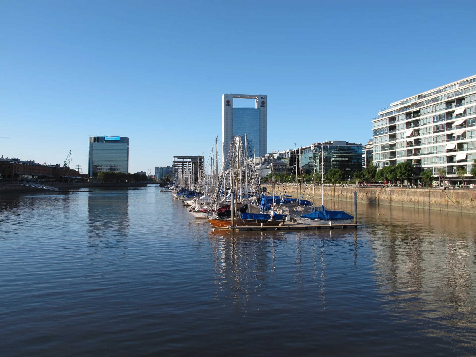 Dique de Puerto Madero em tarde de céu azul, com barcos atracados, água calma e edifícios contemporâneos ao fundo.