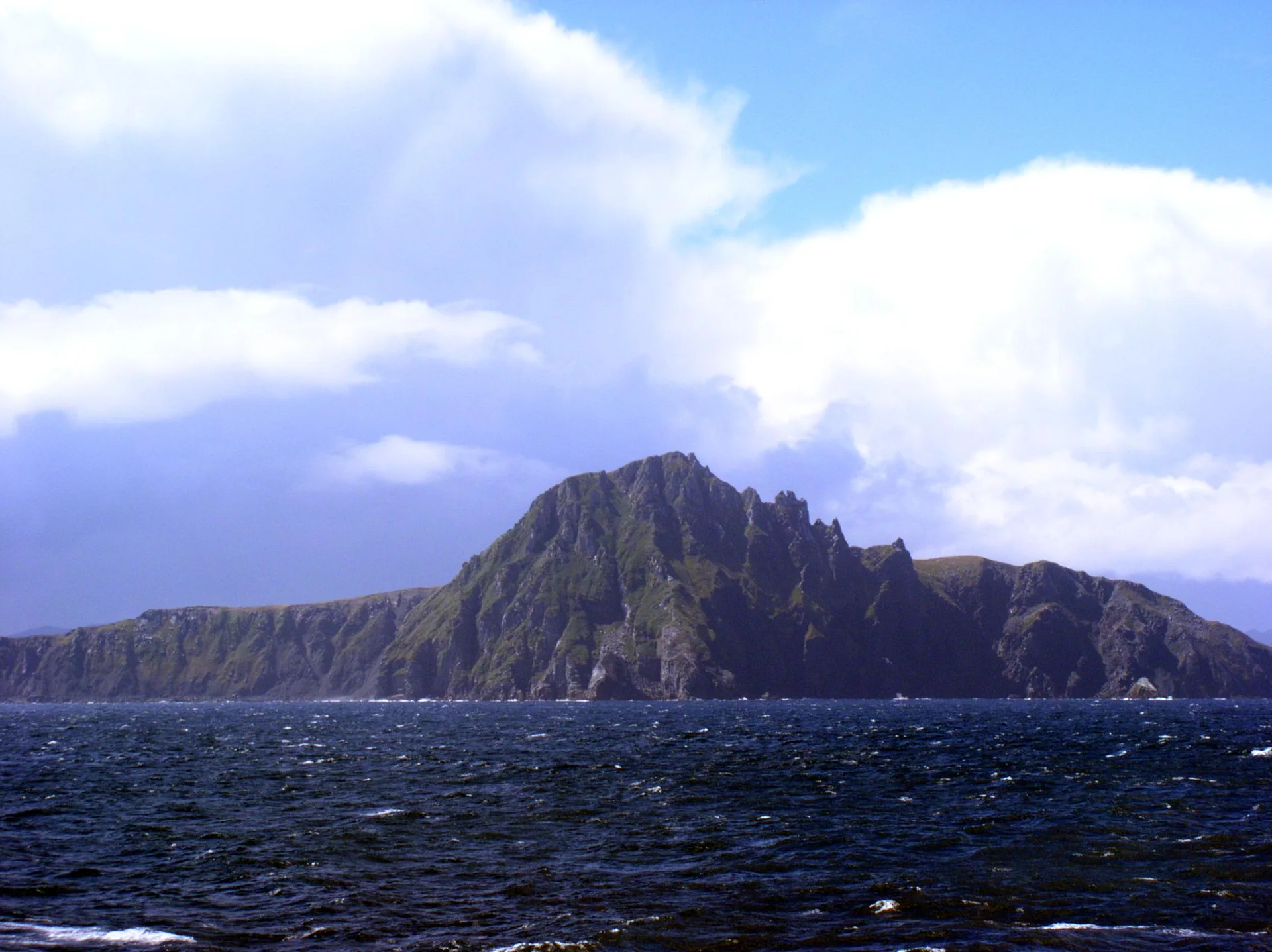 Promontório rochoso do Cabo de Hornos visto do mar, com ondas escuras em primeiro plano e nuvens claras sobre a ilha.